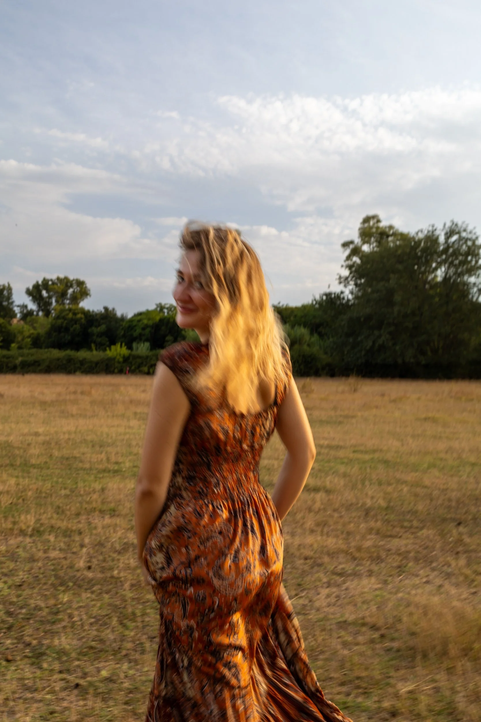 A woman in a colorful dress standing outdoors on a grassy field with trees in the background under a partly cloudy sky, smiling and looking over her shoulder.