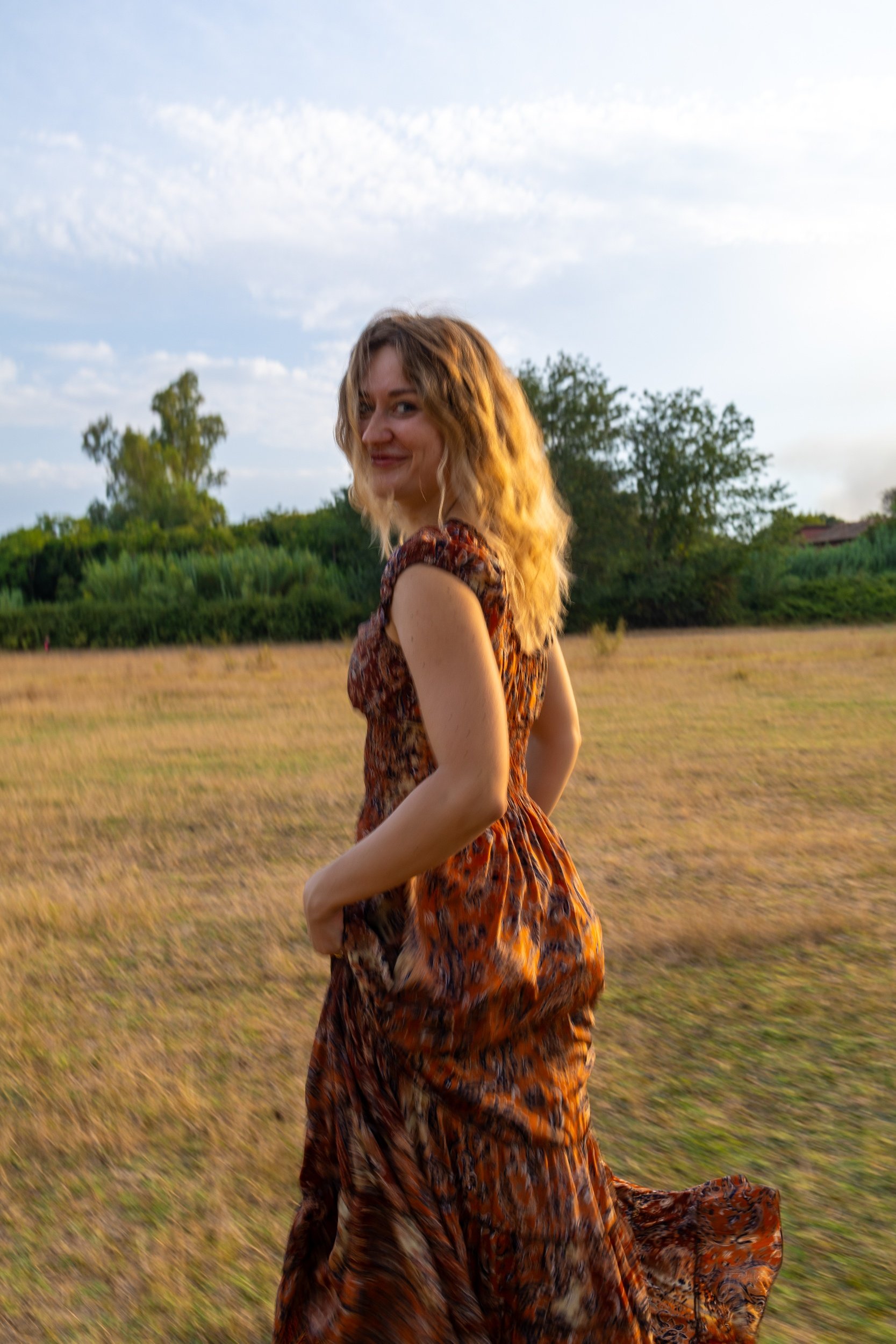 A woman with wavy blonde hair wearing a brown and orange animal print dress standing outdoors in a field with green trees in the background, smiling at the camera during sunset.