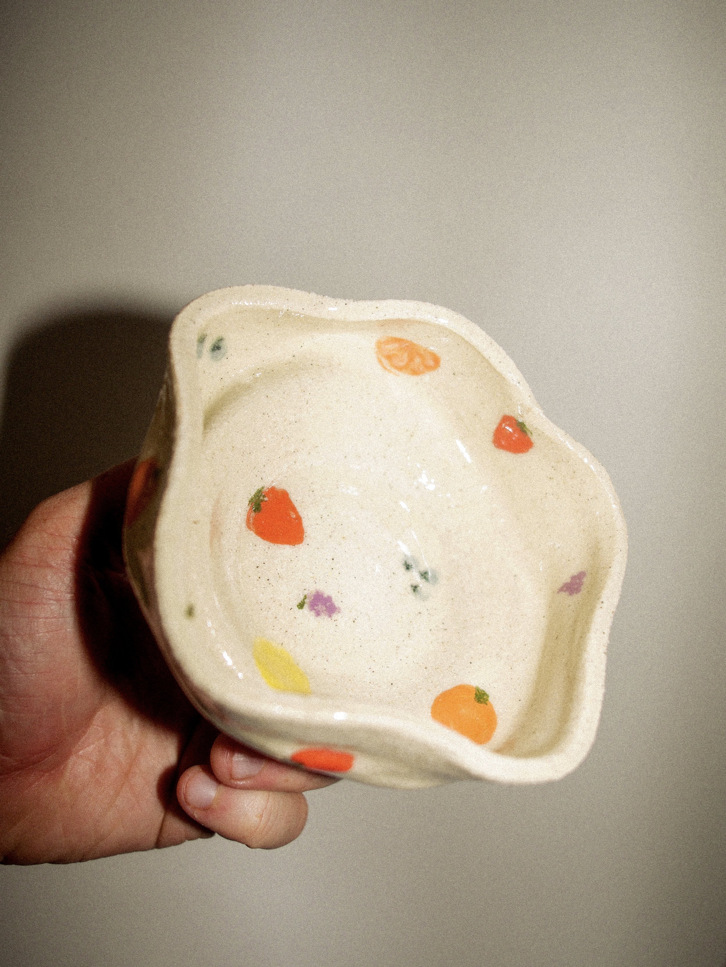 Hand holding a ceramic bowl with colorful fruit and vegetable designs, such as strawberries, carrots, and other small fruits.