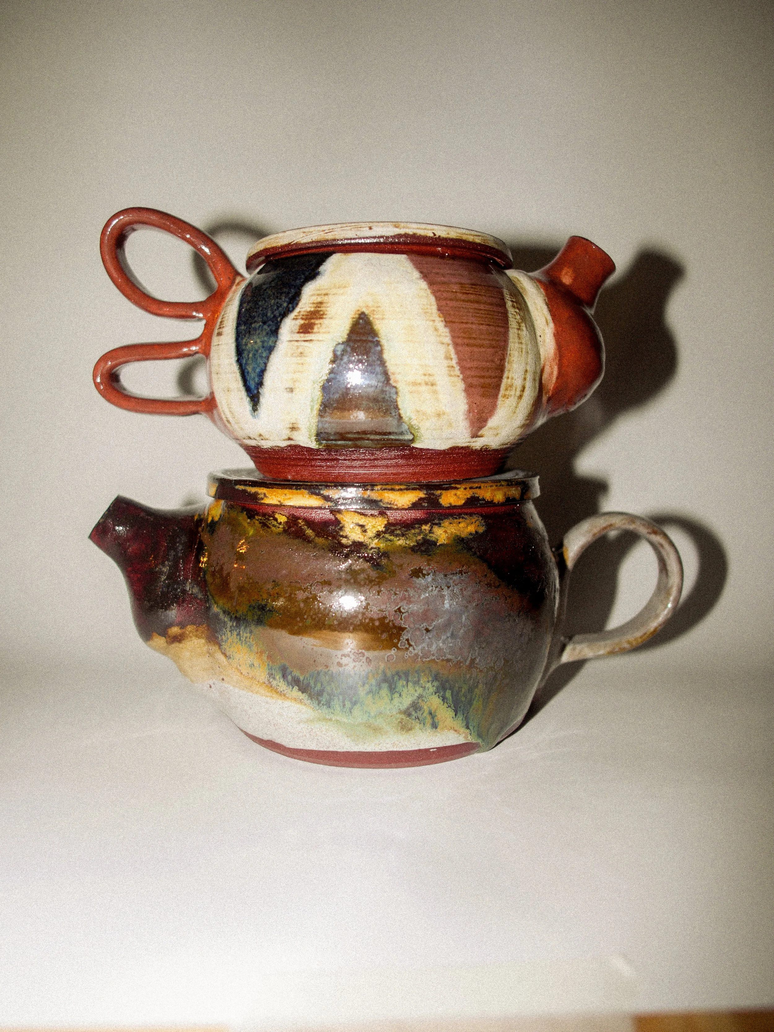 Two ceramic teapots with abstract painted designs stacked on top of each other on a white surface.