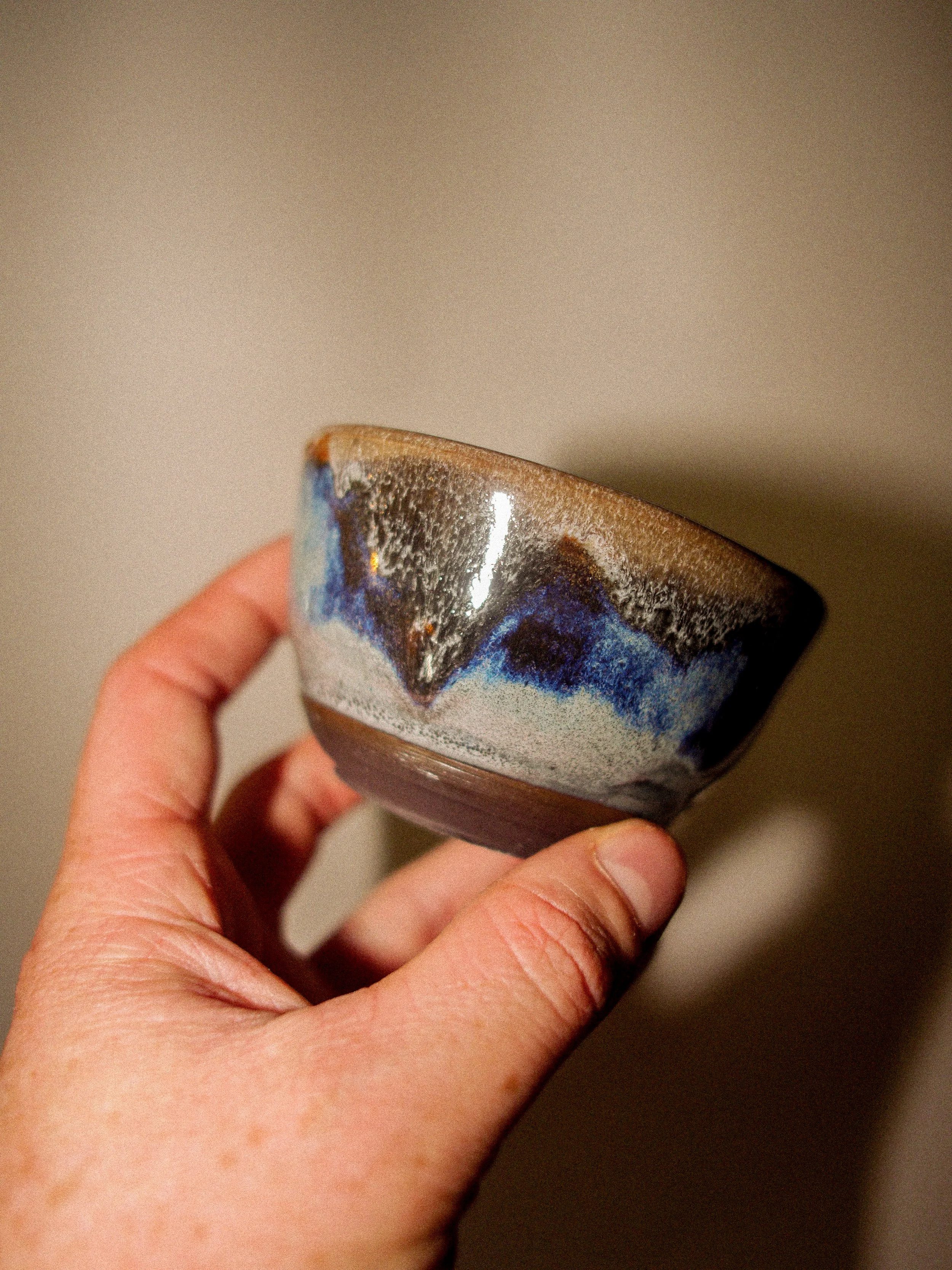 Close-up of a hand holding a small ceramic bowl with a layered, abstract glaze design in shades of brown, black, blue, and white.