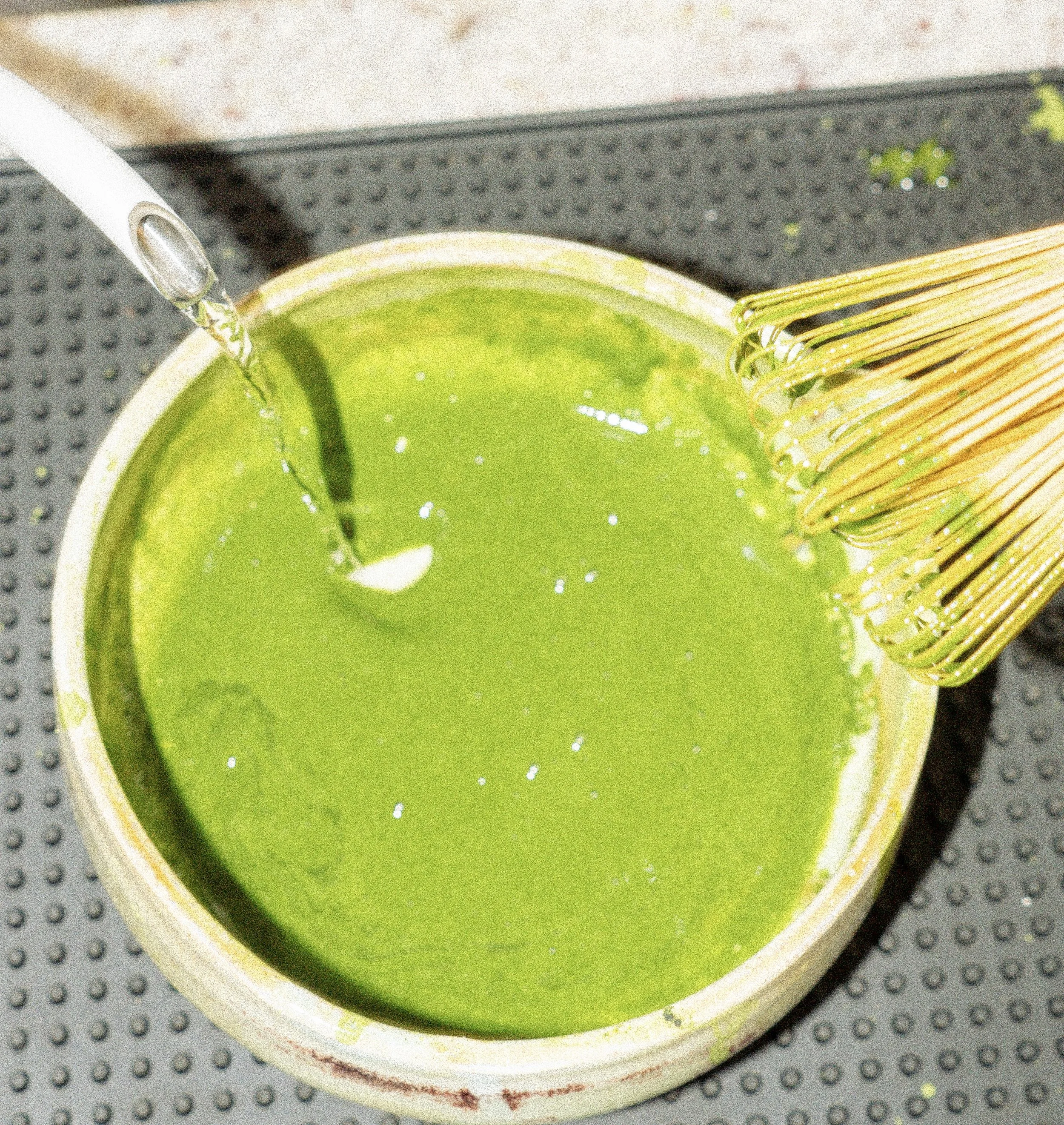 A container with green matcha tea being prepared, with a whisk on the side and water being poured into the mixture.