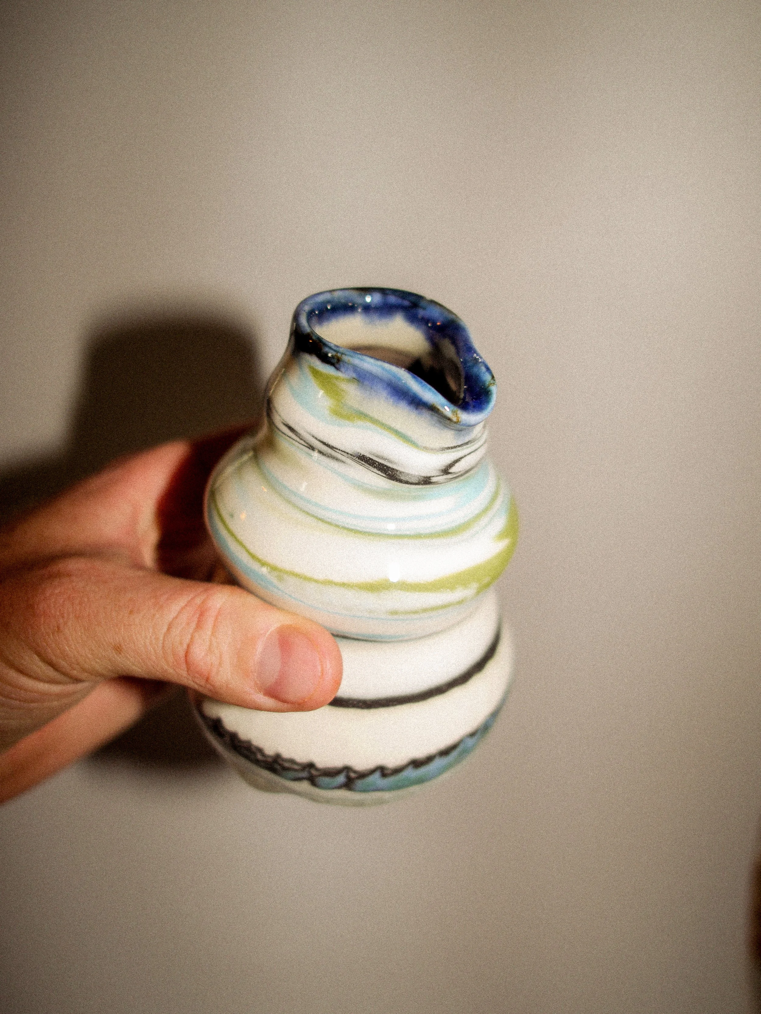 A small, colorful ceramic vase with a wavy opening held by a person's hand, set against a plain background.