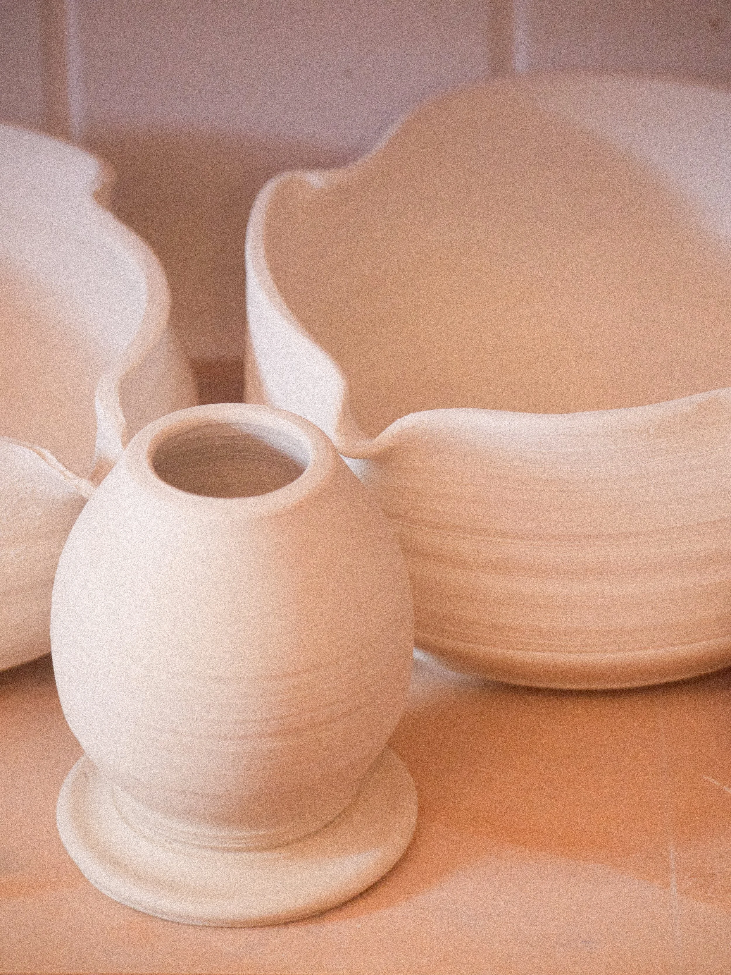 Several cream-colored, unglazed pottery pieces, including a small vase on a matching saucer, and large, petal-shaped bowls stacked together, displayed on a light wooden surface.