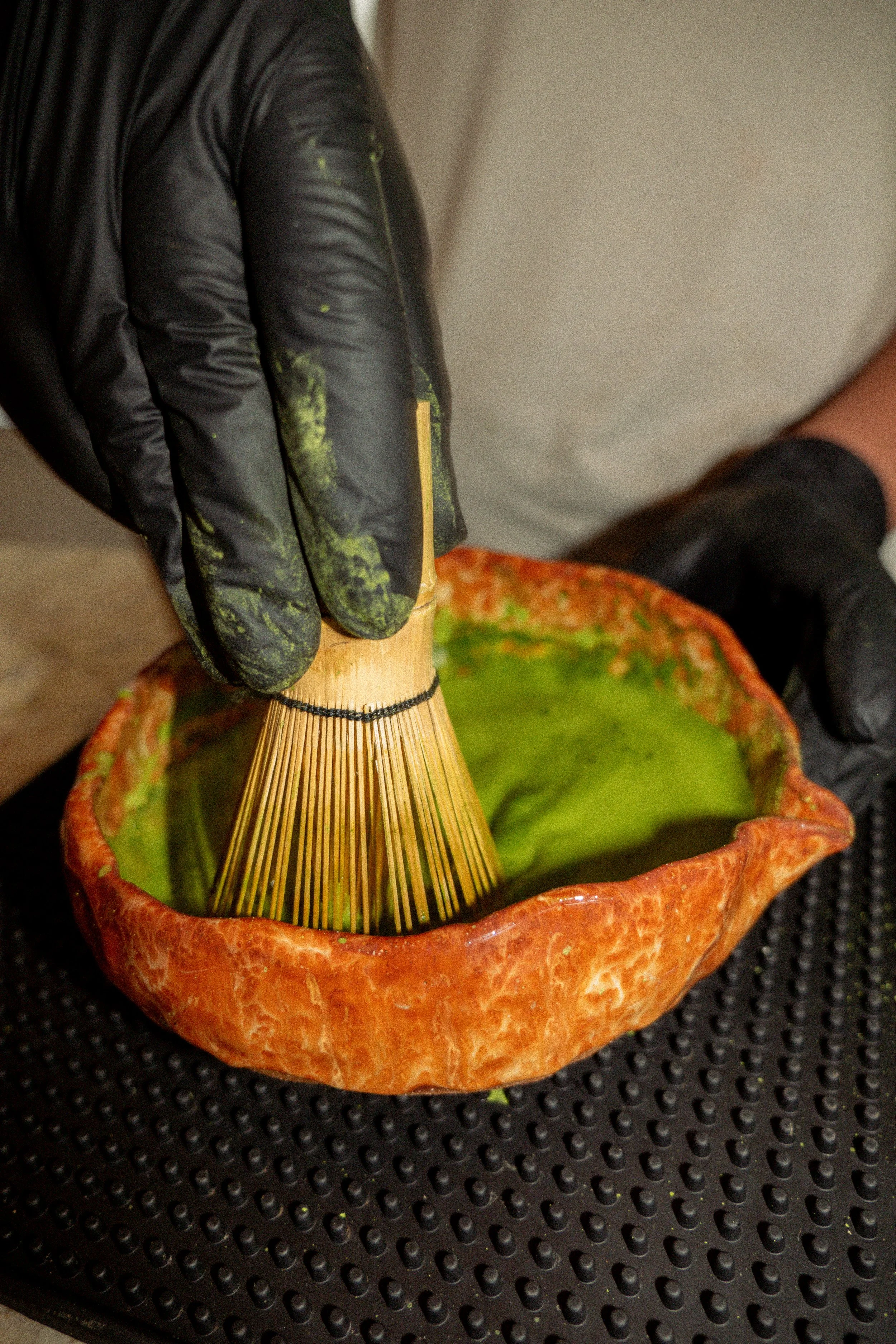 Person wearing black gloves whisking matcha green tea in a rustic, orange ceramic bowl.