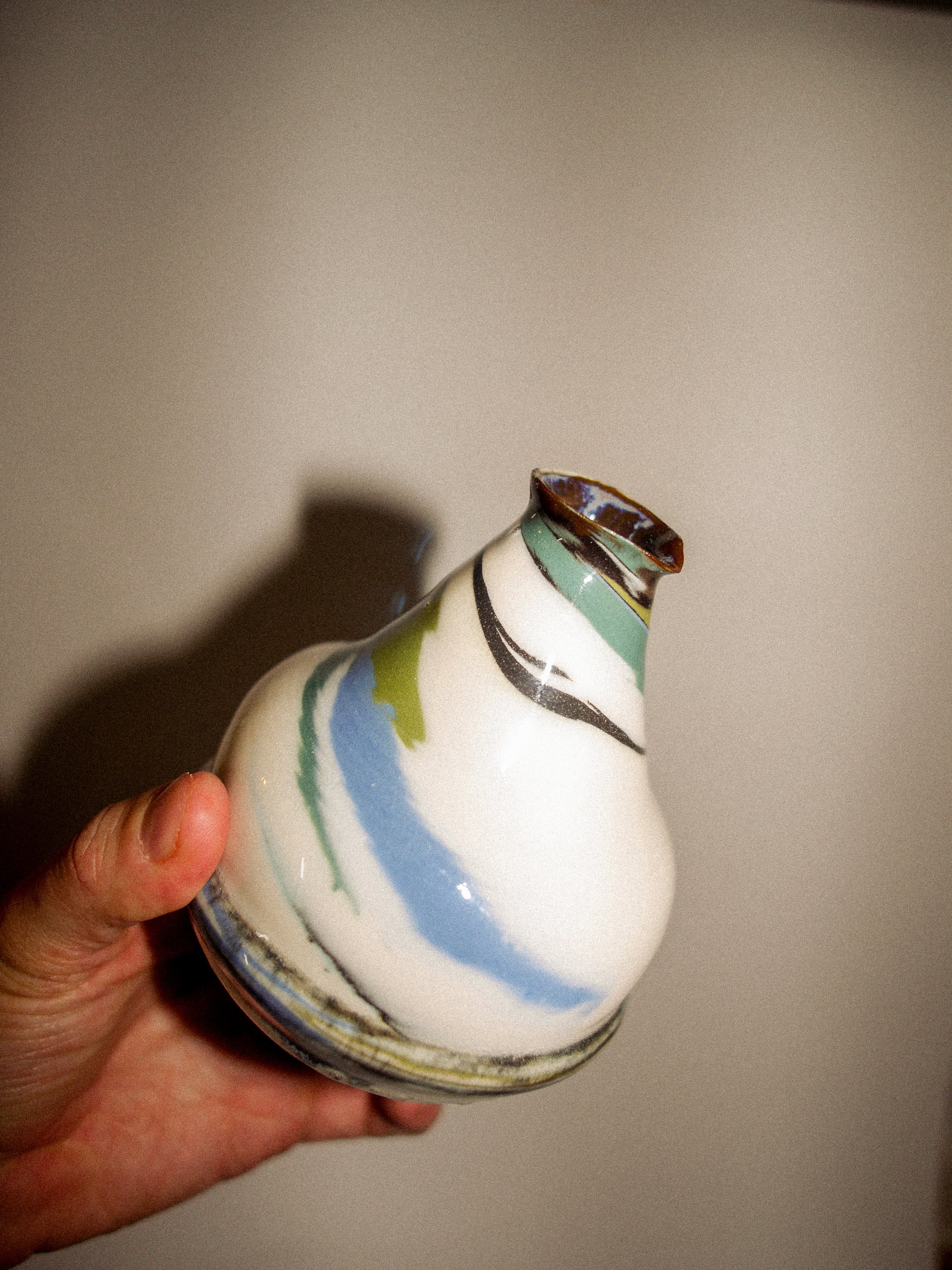 Hand holding a ceramic vase with abstract green, blue, black, and brown brush strokes, set against a plain beige background.