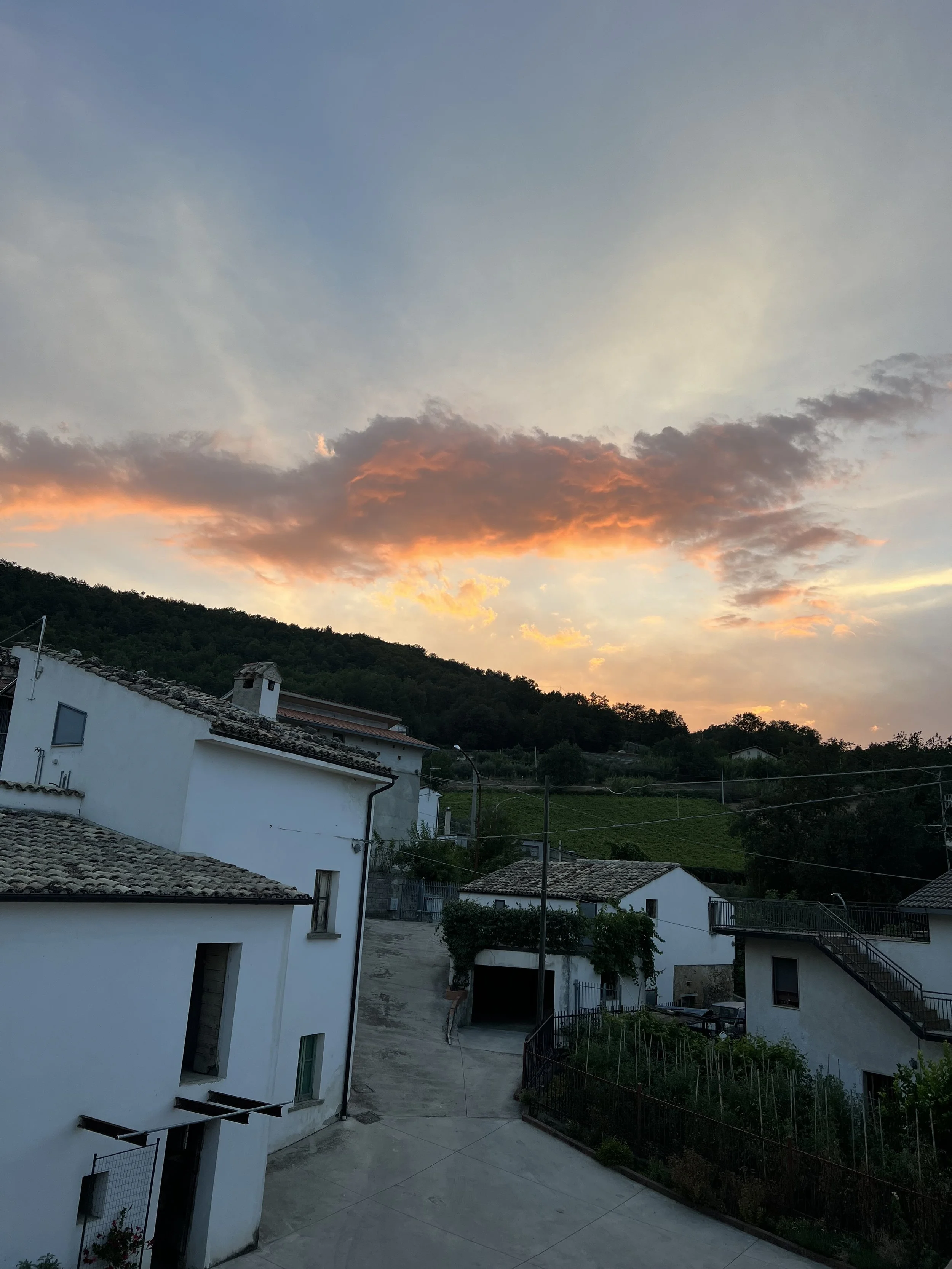 A view of a quiet countryside with white houses, a narrow road, and a hillside covered in greenery under a colorful sky during sunset.