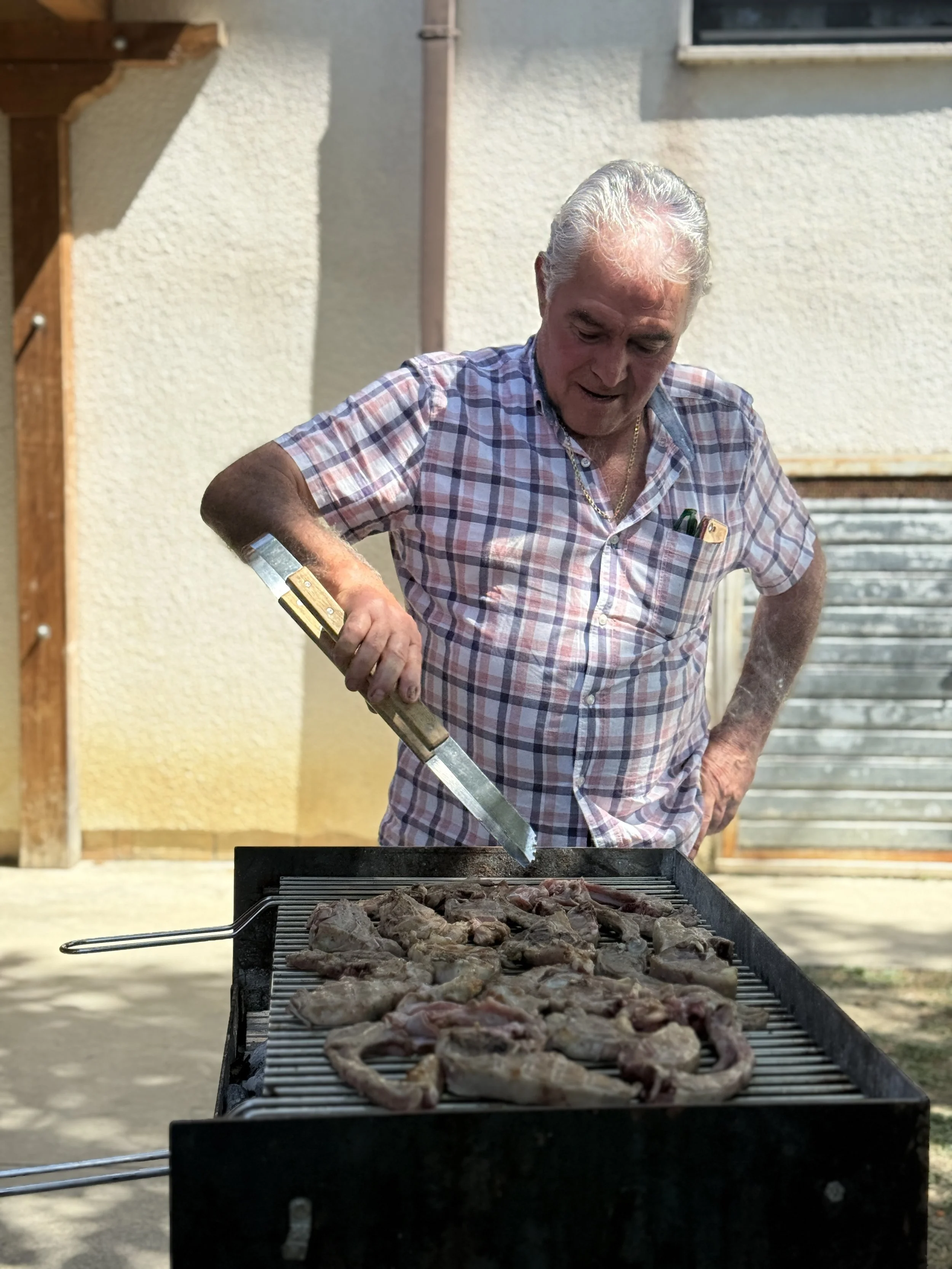 An elderly man wearing a checkered shirt barbequing meat on a grill outdoors.