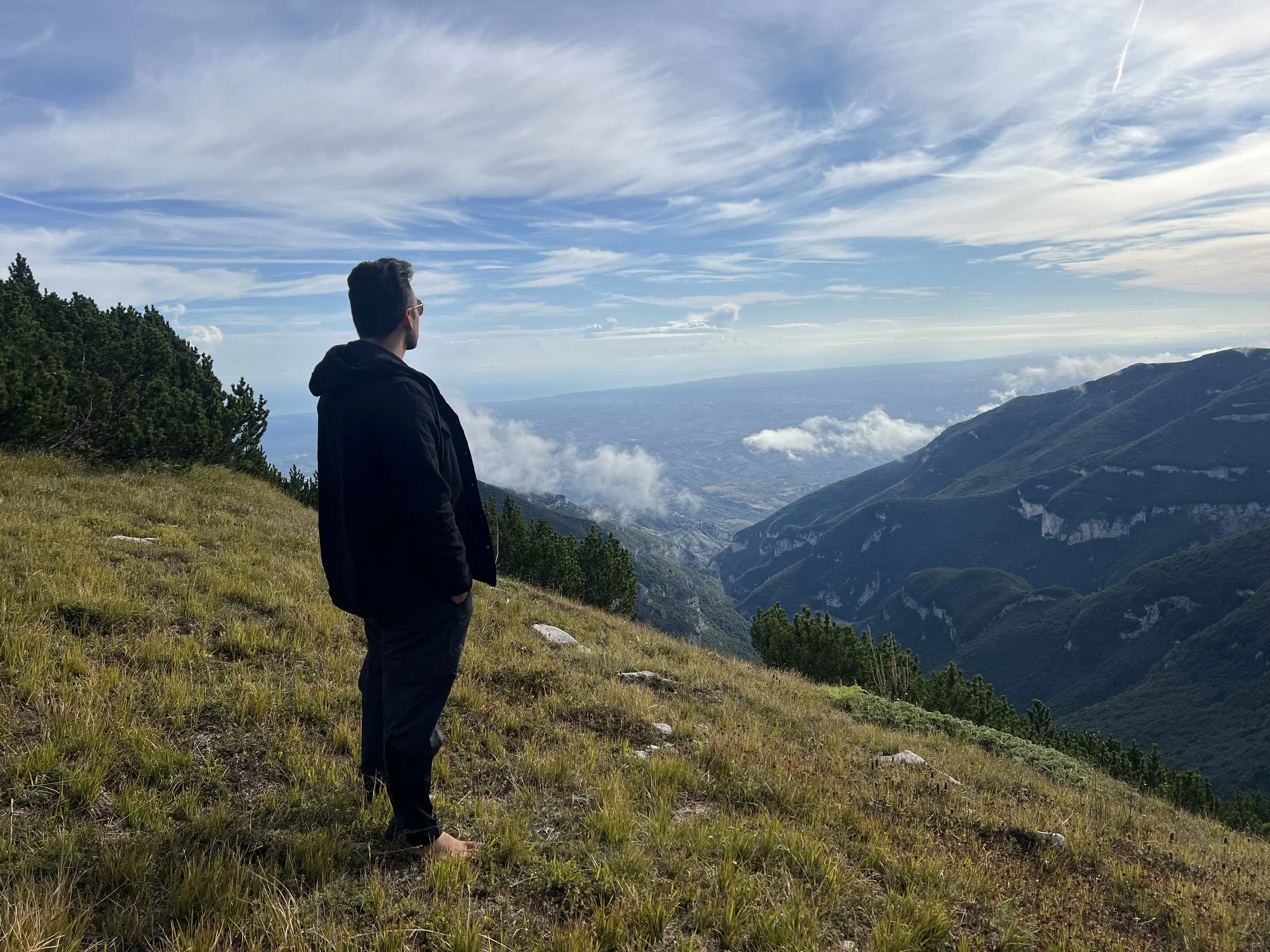 Man in black jacket and pants standing barefoot on grassy hillside, looking at mountain view with green peaks and clouds in the sky.