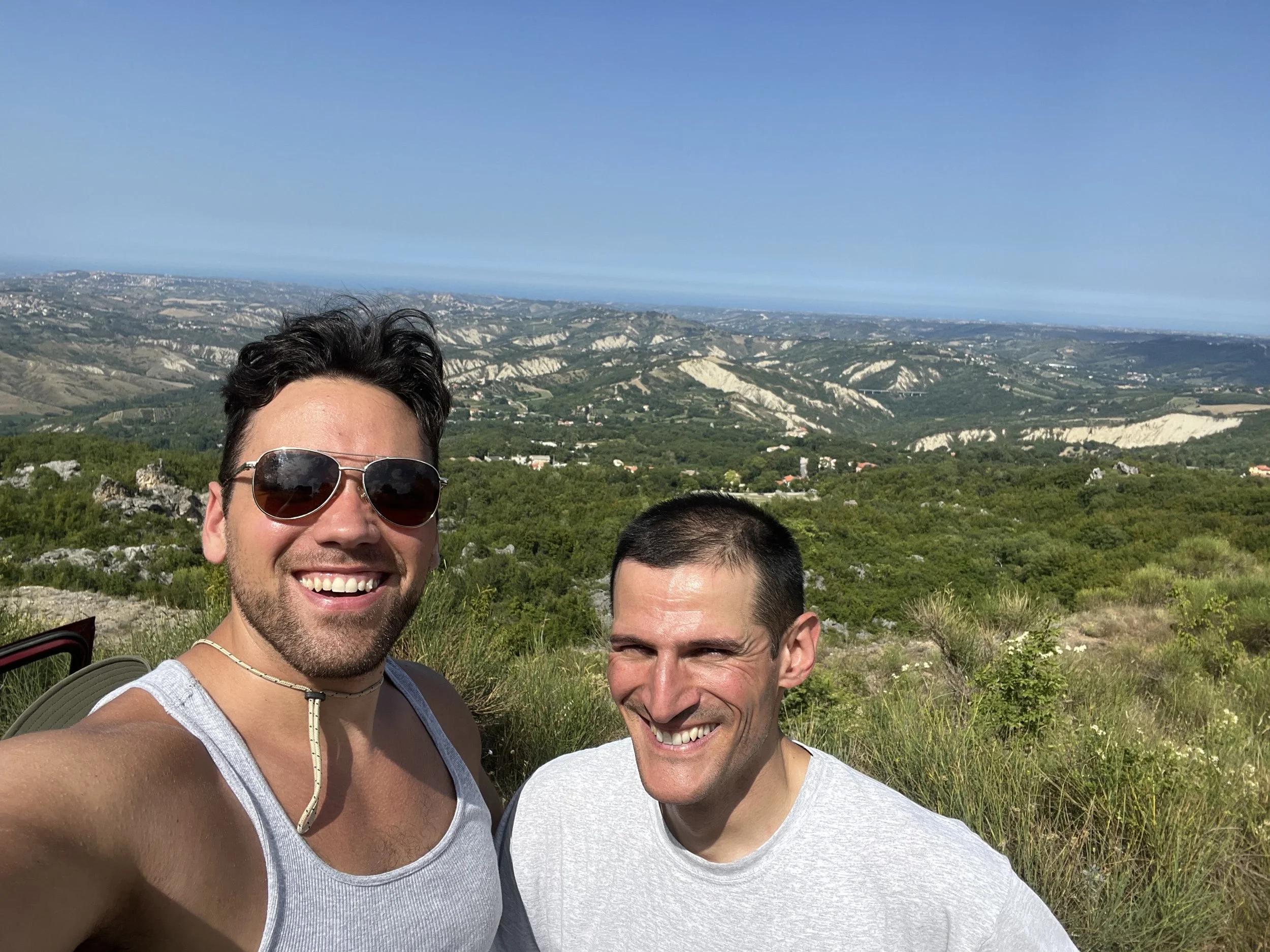 Two smiling men taking a selfie outdoors with a scenic landscape of rolling hills, greenery, and a clear blue sky in the background.