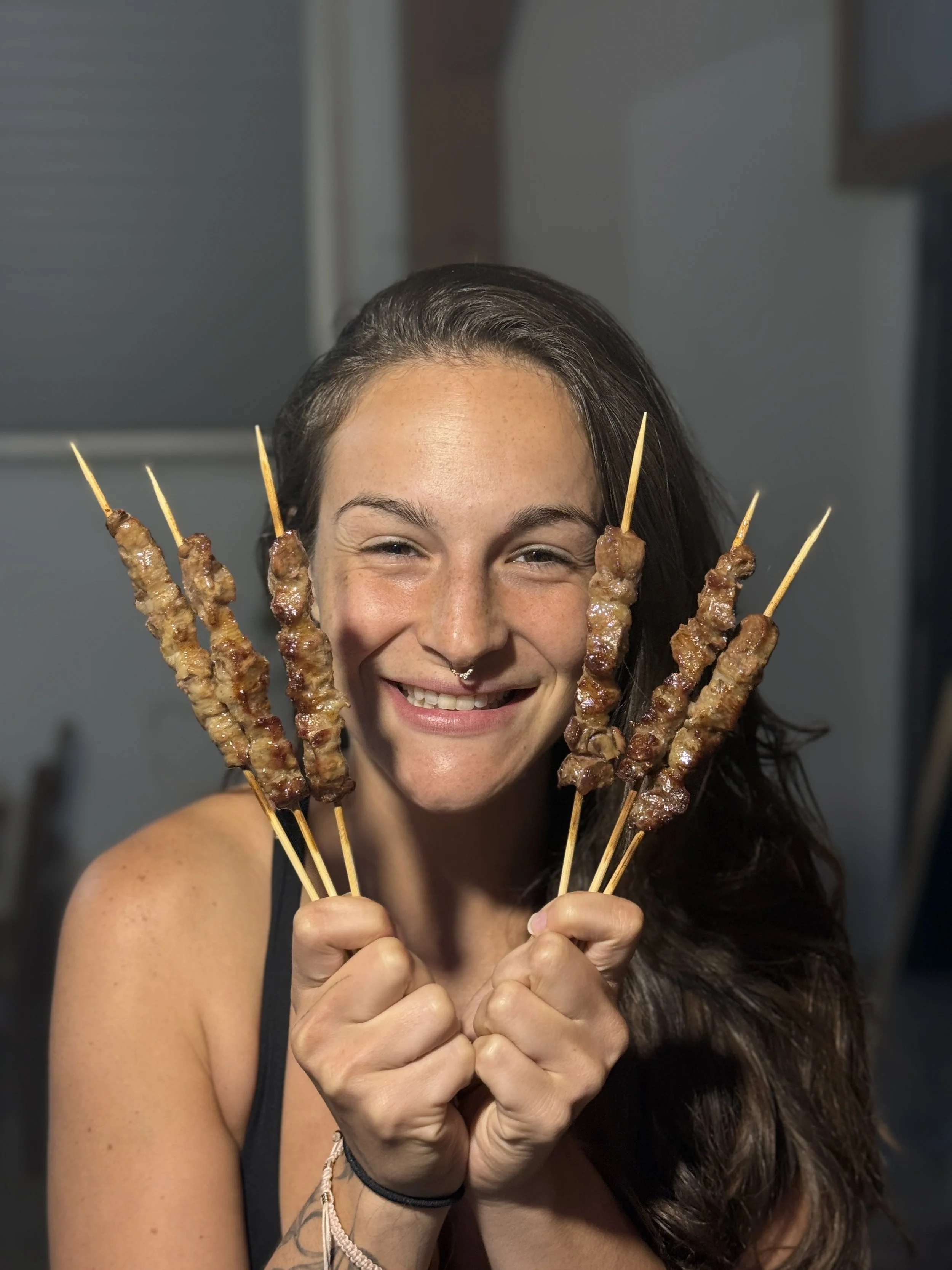 A woman smiling and holding several skewers of cooked meat in each hand.