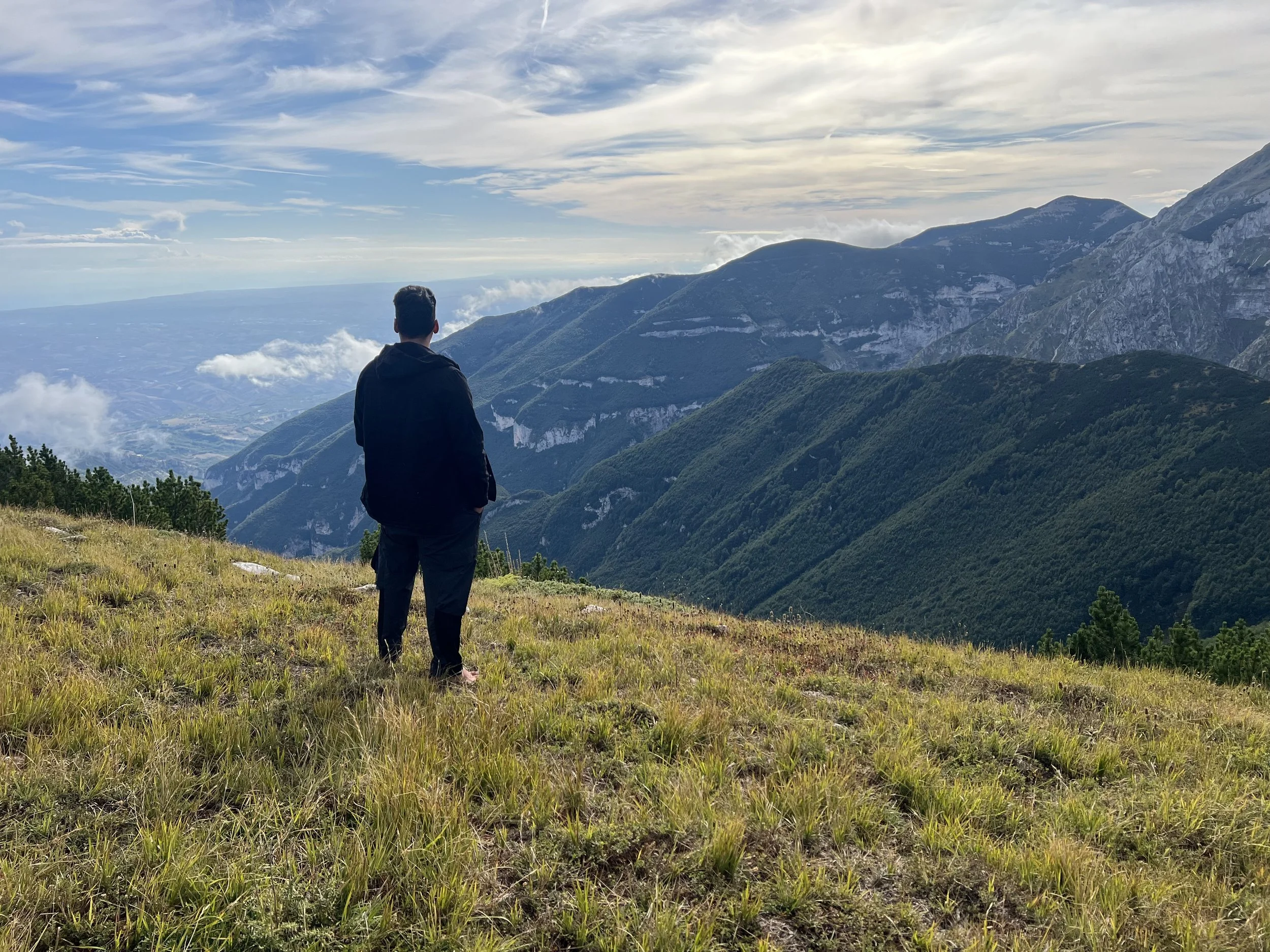 A person with dark hair wearing a black jacket and black pants stands on a grassy hillside overlooking green mountain ranges under a partly cloudy sky.