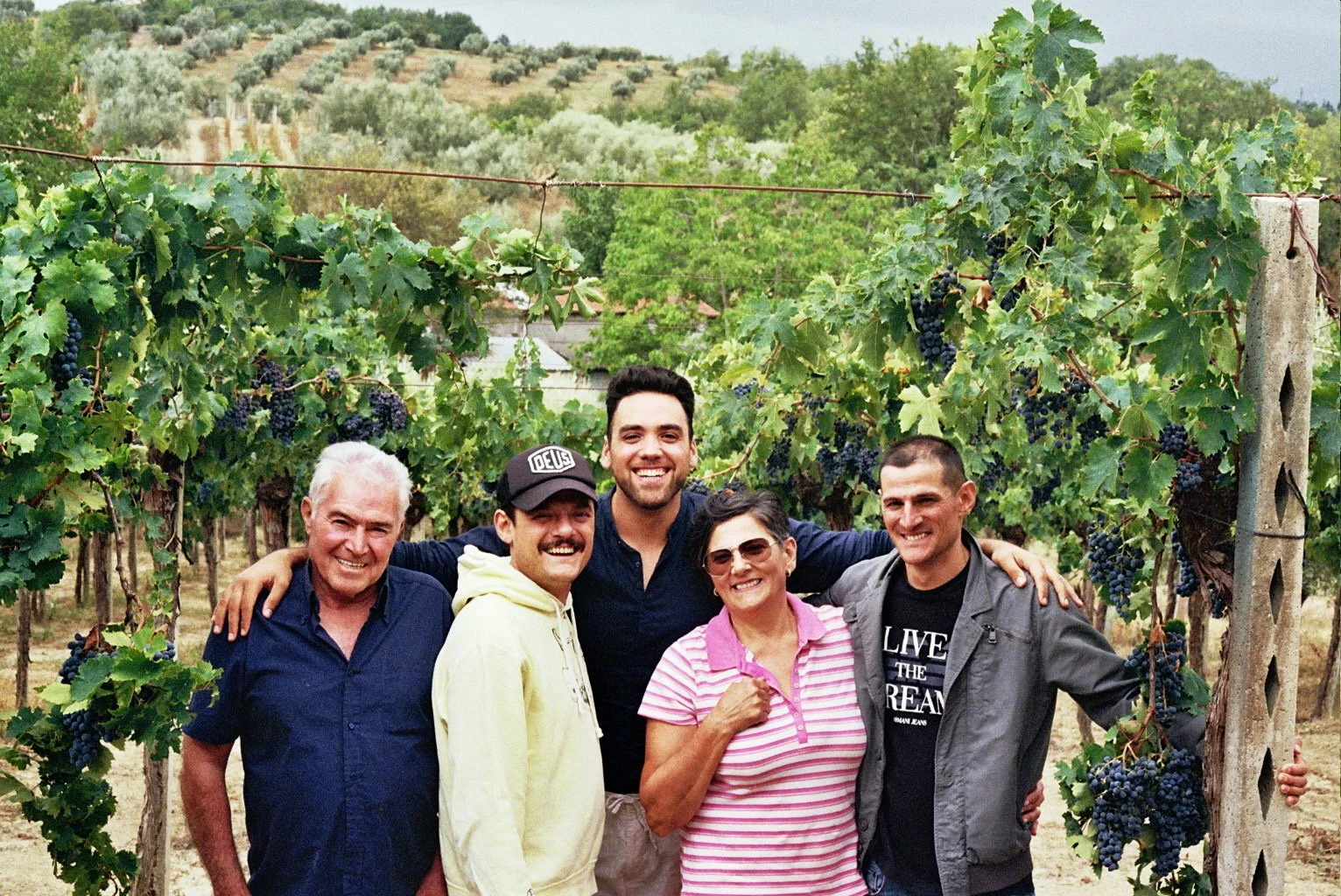 The family of Soul of Abruzzo and the founder smiling and standing together in a vineyard with grapevines and clusters of grapes, with hills in the background.