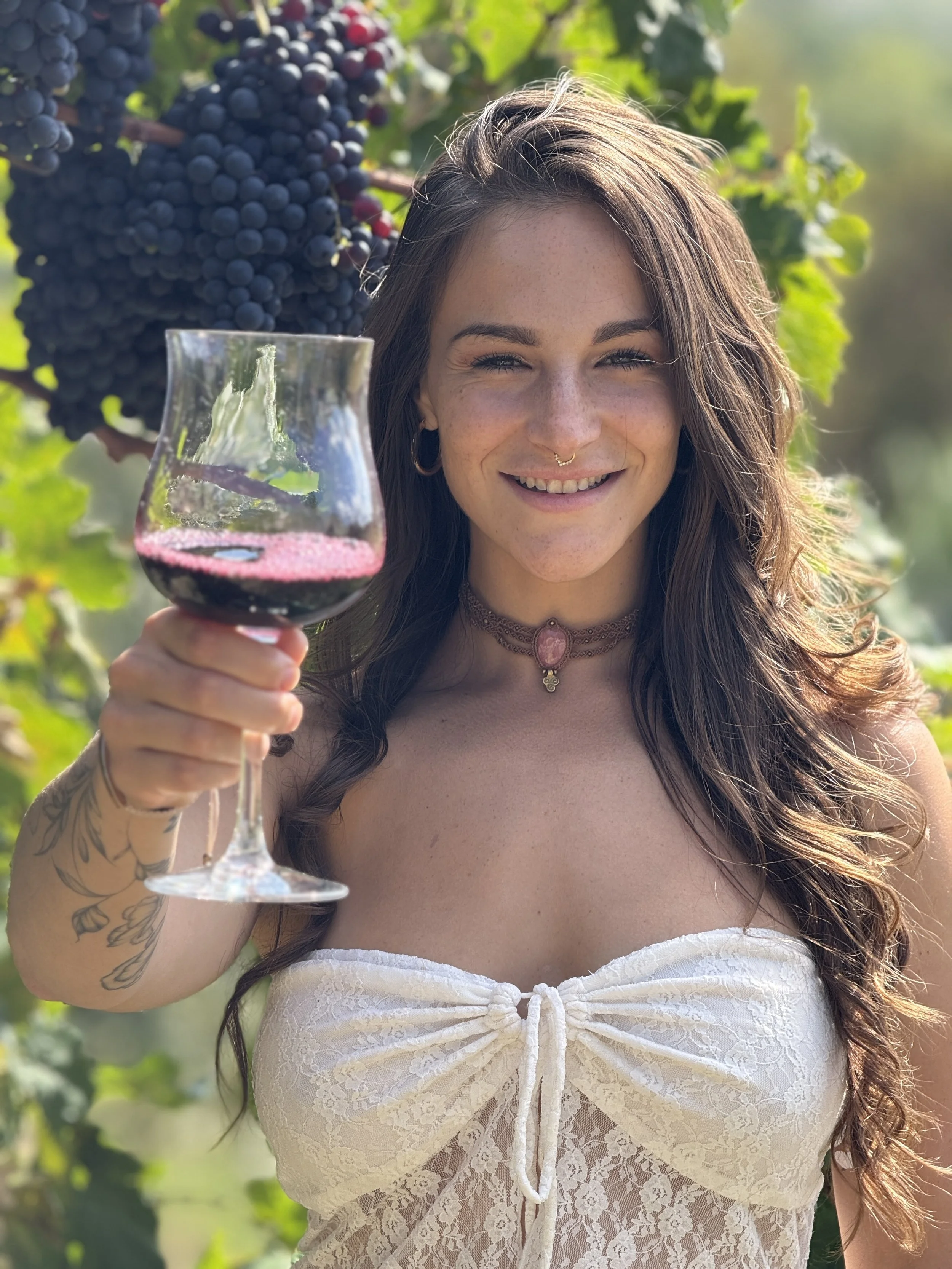 A woman with long brown hair, smiling, holding a glass of red wine outdoors among grapevines, with a cluster of purple grapes hanging in the background.