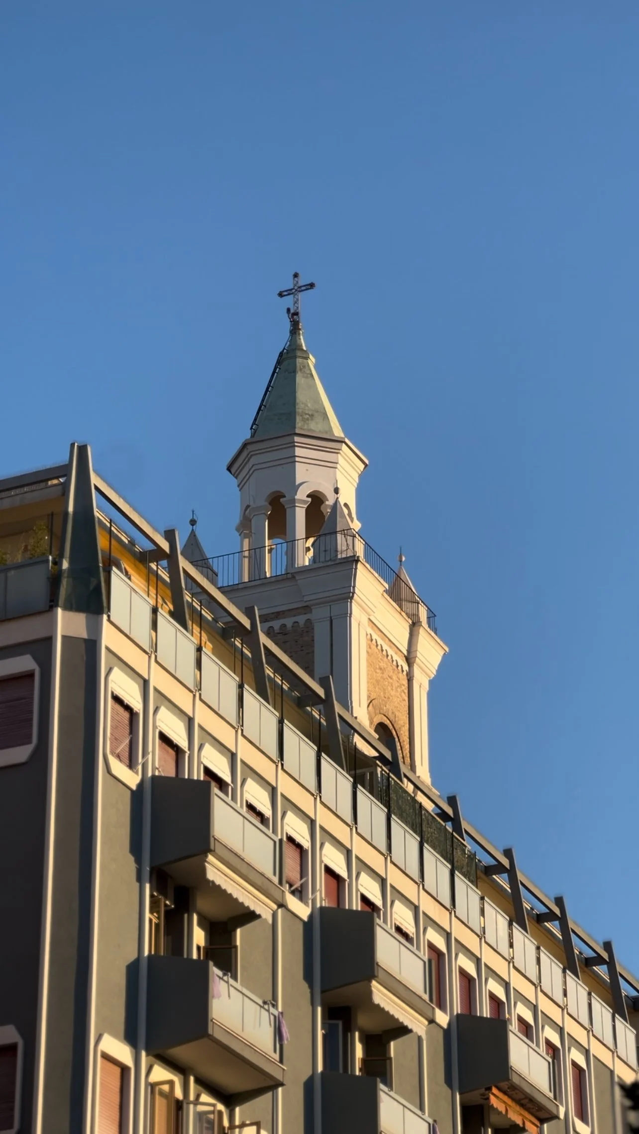 A tall church steeple with a cross on top, seen behind a multi-story residential building with balconies, under a clear blue sky.