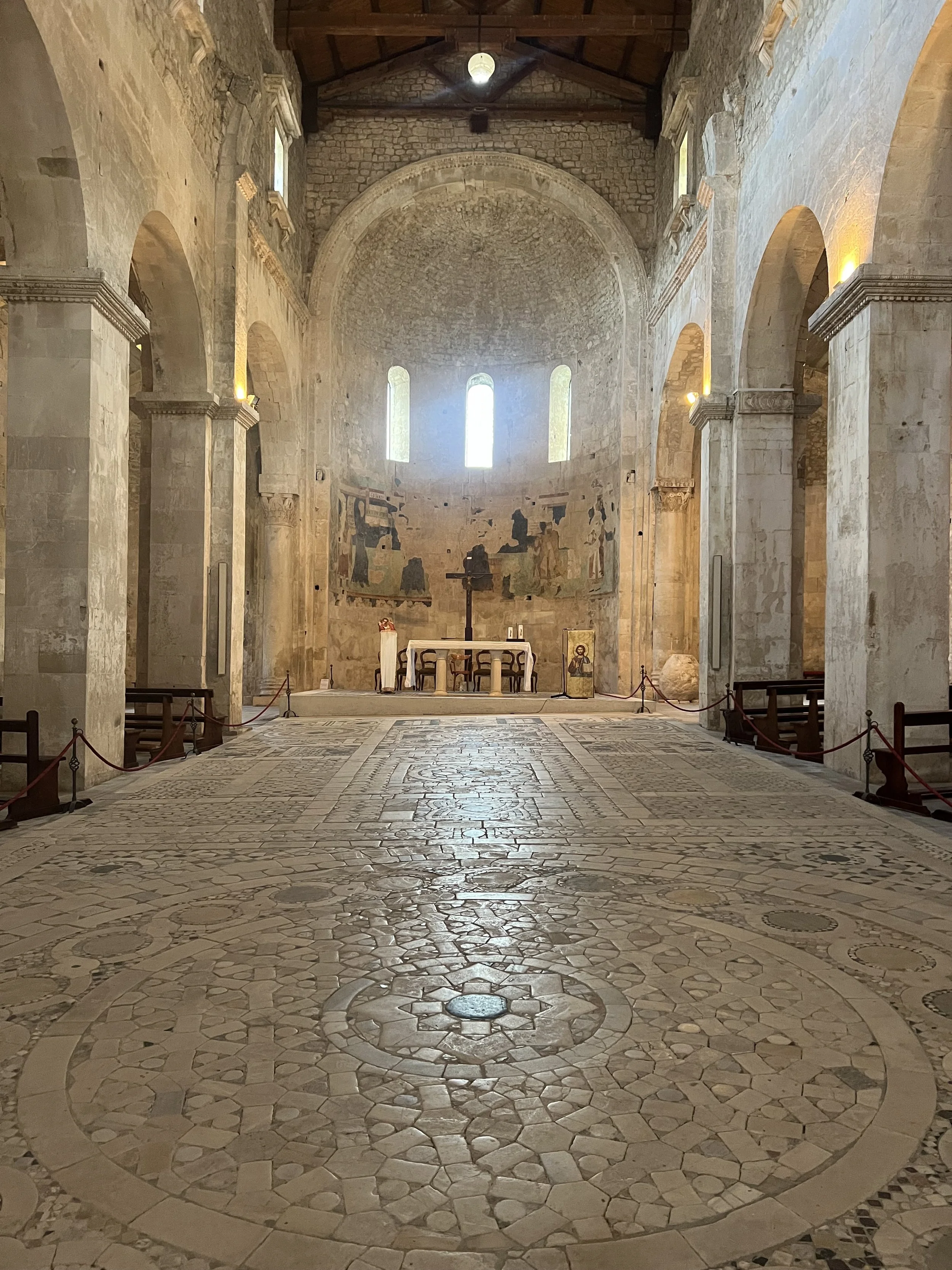 Interior of a historic church with stone walls and floors, featuring an altar with a crucifix, religious icons, and a fresco on the back wall. Sunlight shines through tall, narrow windows.