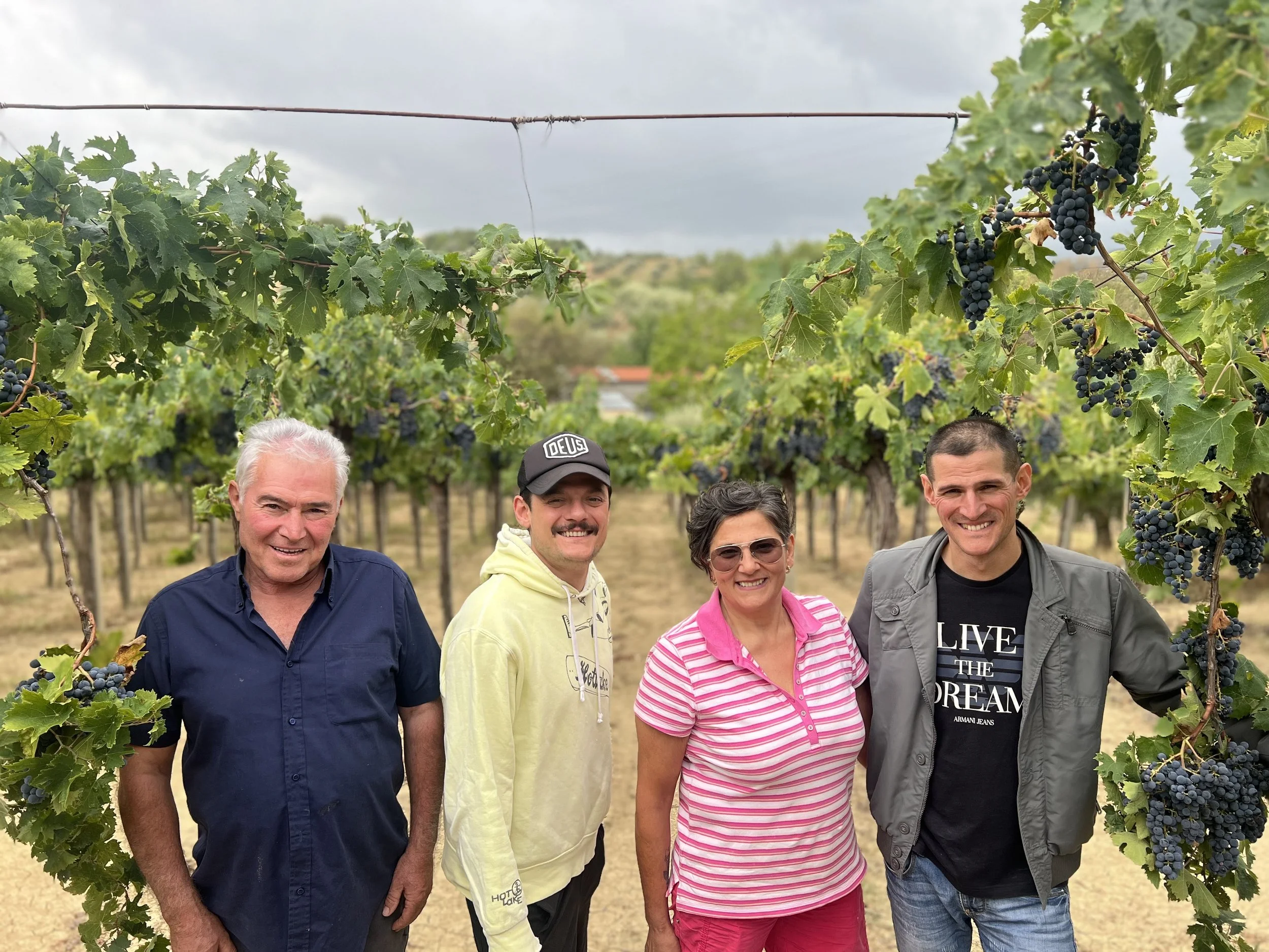 The Soul of Abruzzo standing in a vineyard with grapevines and clusters of dark purple grapes around them, smiling at the camera.