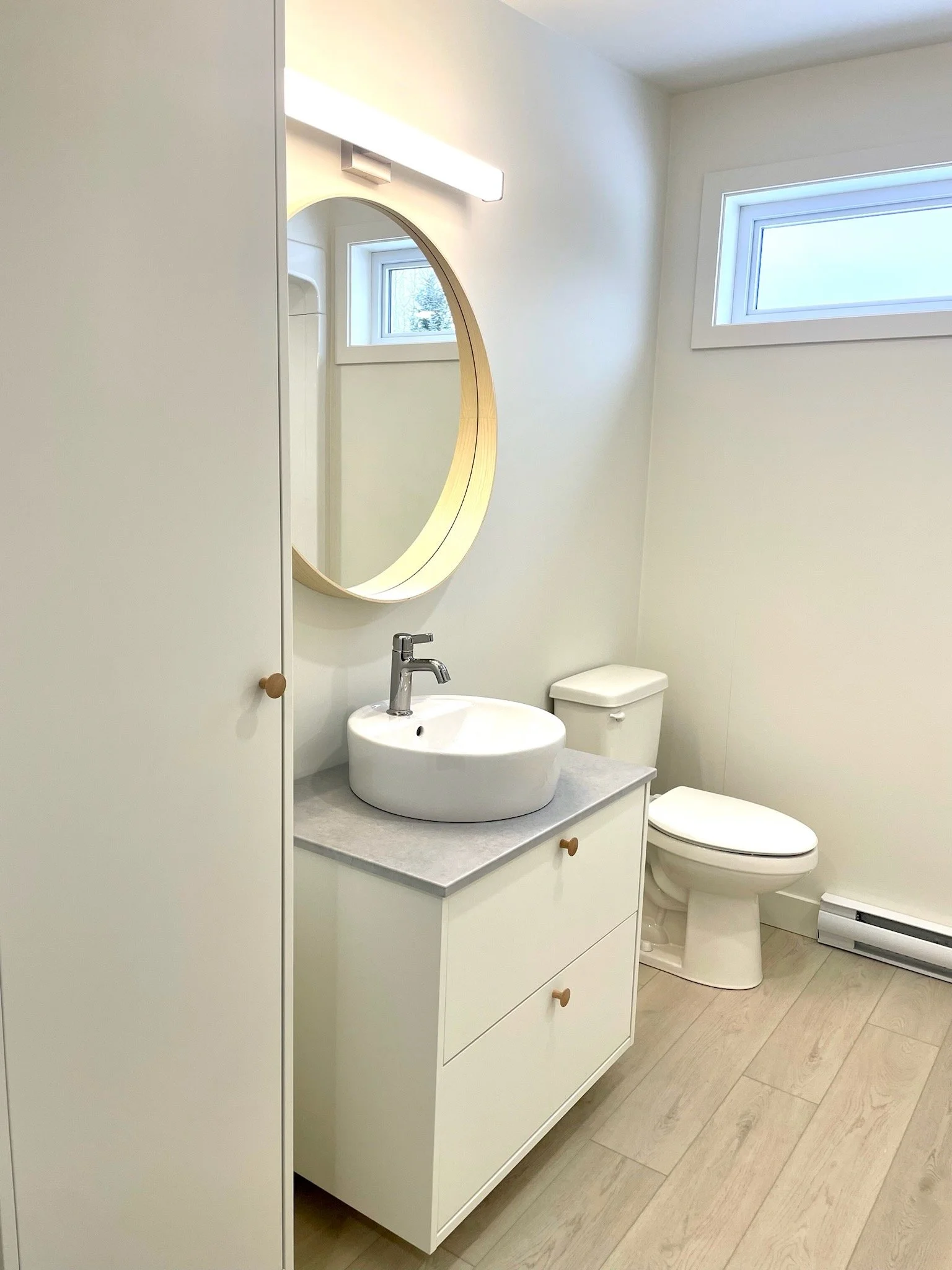 Petit toilette dans une salle de bain avec un lavabo rond, un miroir oval, une toilette et deux fenêtres en haut.