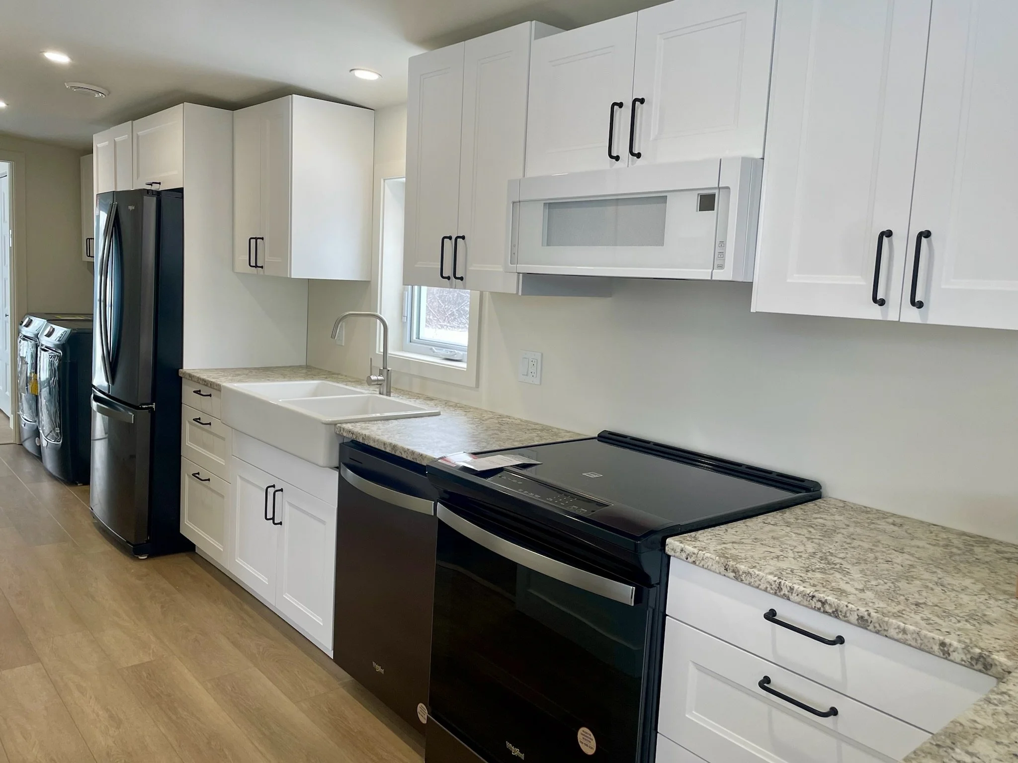 Cuisine moderne avec armoires blanches, comptoir en granite, évier double en céramique, micro-ondes encastré, réfrigérateur noir, lave-vaisselle noir, cuisinière électrique et sol en bois.