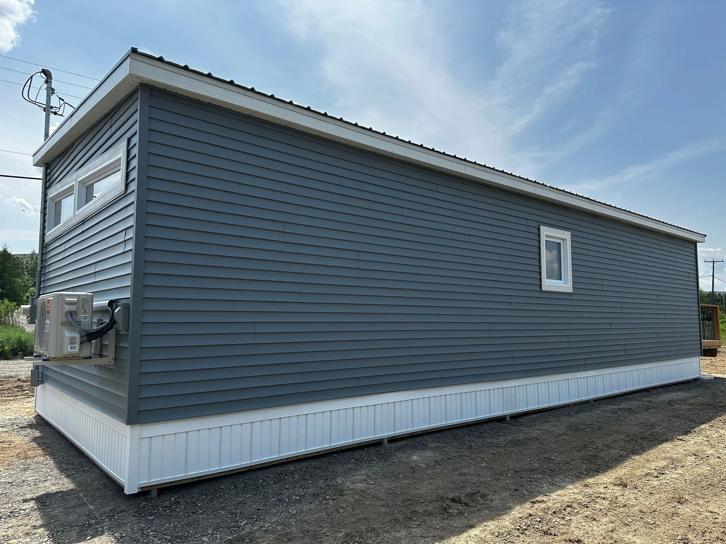 Maison mobile avec siding bleu, deux fenêtres et unité de climatisation extérieure, sous un ciel en partie nuageux.