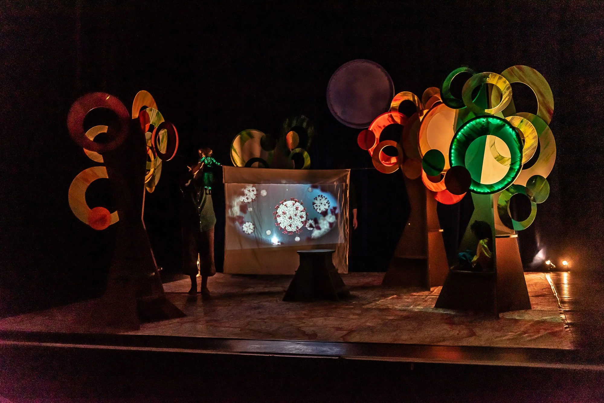 Dark stage with large colorful, abstract tree art installations on each side, a projection of virus images on a screen, and people partially visible in the background, illuminated by ambient lights.