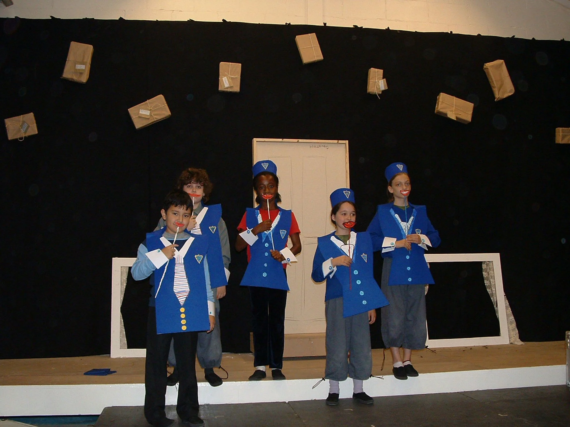 Children dressed as characters in a play, wearing blue and white costumes with striped ties, on stage with black background and paper packages hanging above.