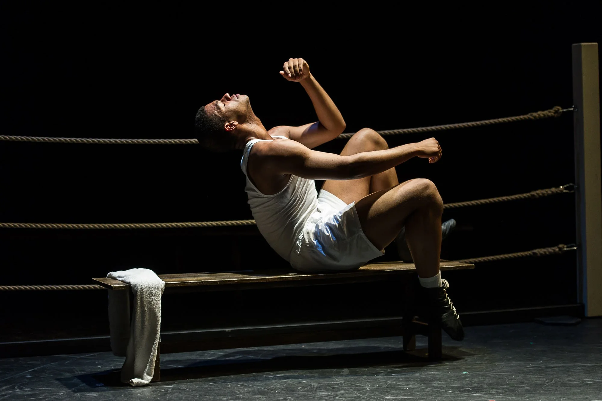 A male boxer in white athletic clothing and boxing shoes sitting on a bench in a boxing ring, leaning back with his head tilted up and one arm bent, surrounded by dark space and boxing ropes.