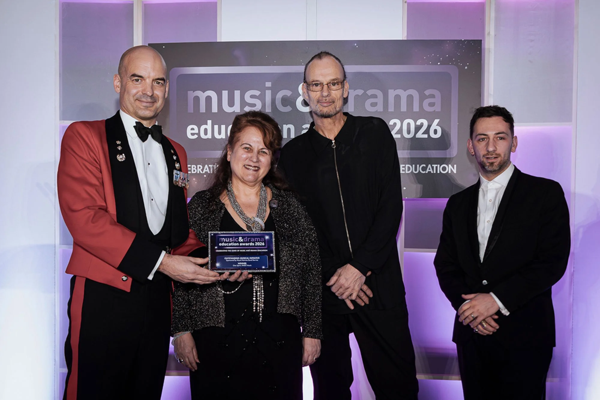 The One Spirit Team standing together at the awards event, holding the plaque, with a backdrop that reads 'music & drama education awards 2026'