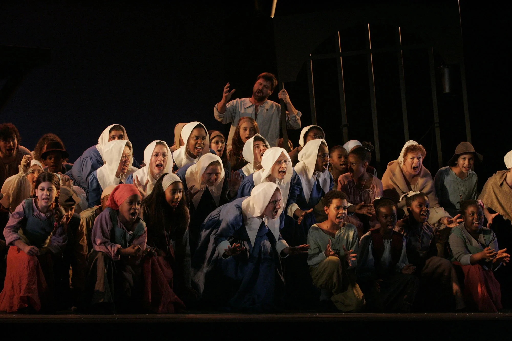 A theatrical scene with children in period costumes portraying nuns, peasants, and other roles on stage with a dark background, showing expressions of anger, fear, or sadness, and a man standing behind them with his hands raised.