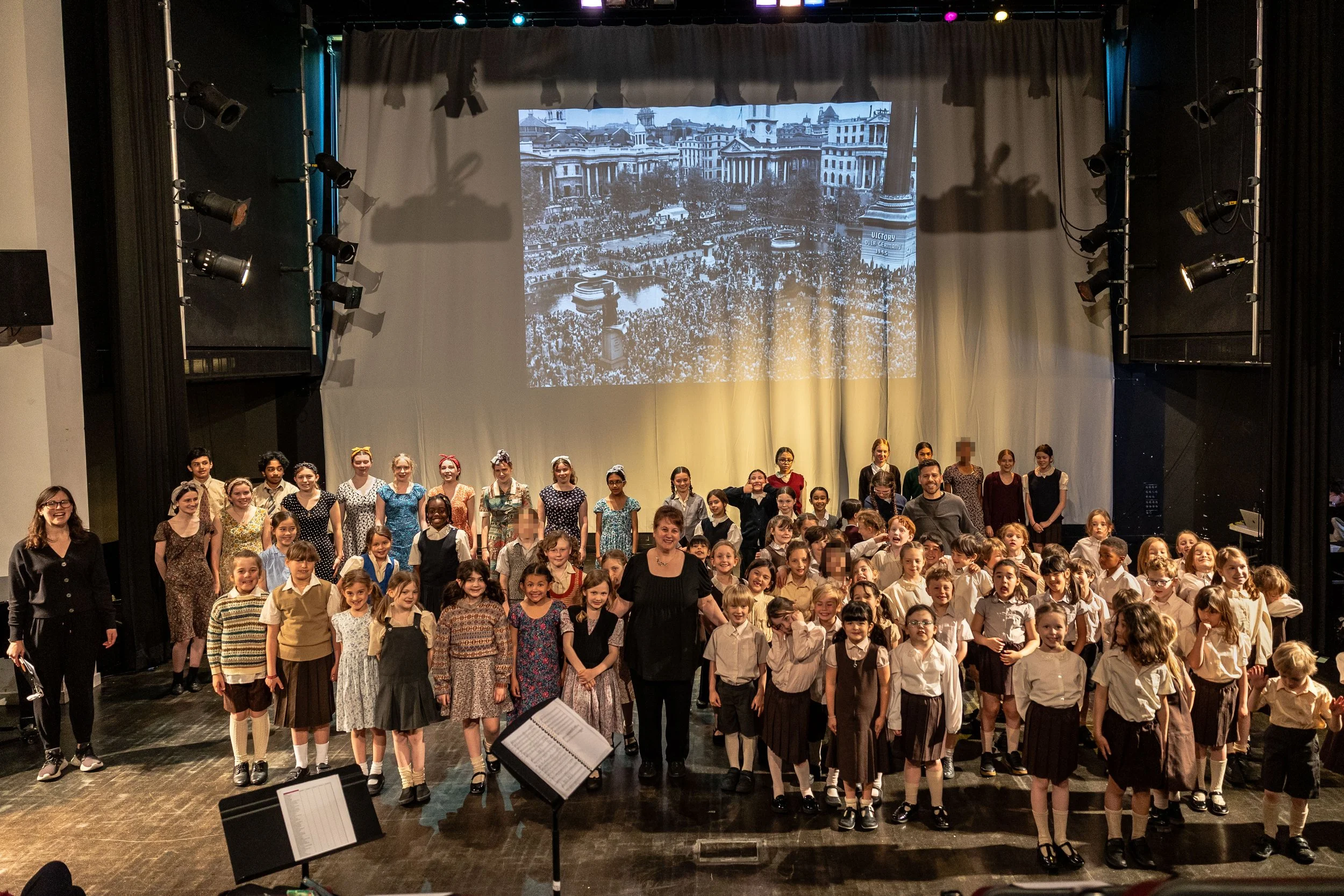 A large group of children and a few adults gathered on a stage for a performance or photograph, with a backdrop showing a historical black and white photo of a city square with buildings and a crowd.