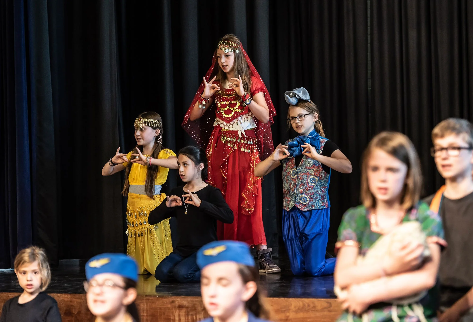 Children performing on stage in colorful costumes, with some kneeling and some standing, during a school play or performance.
