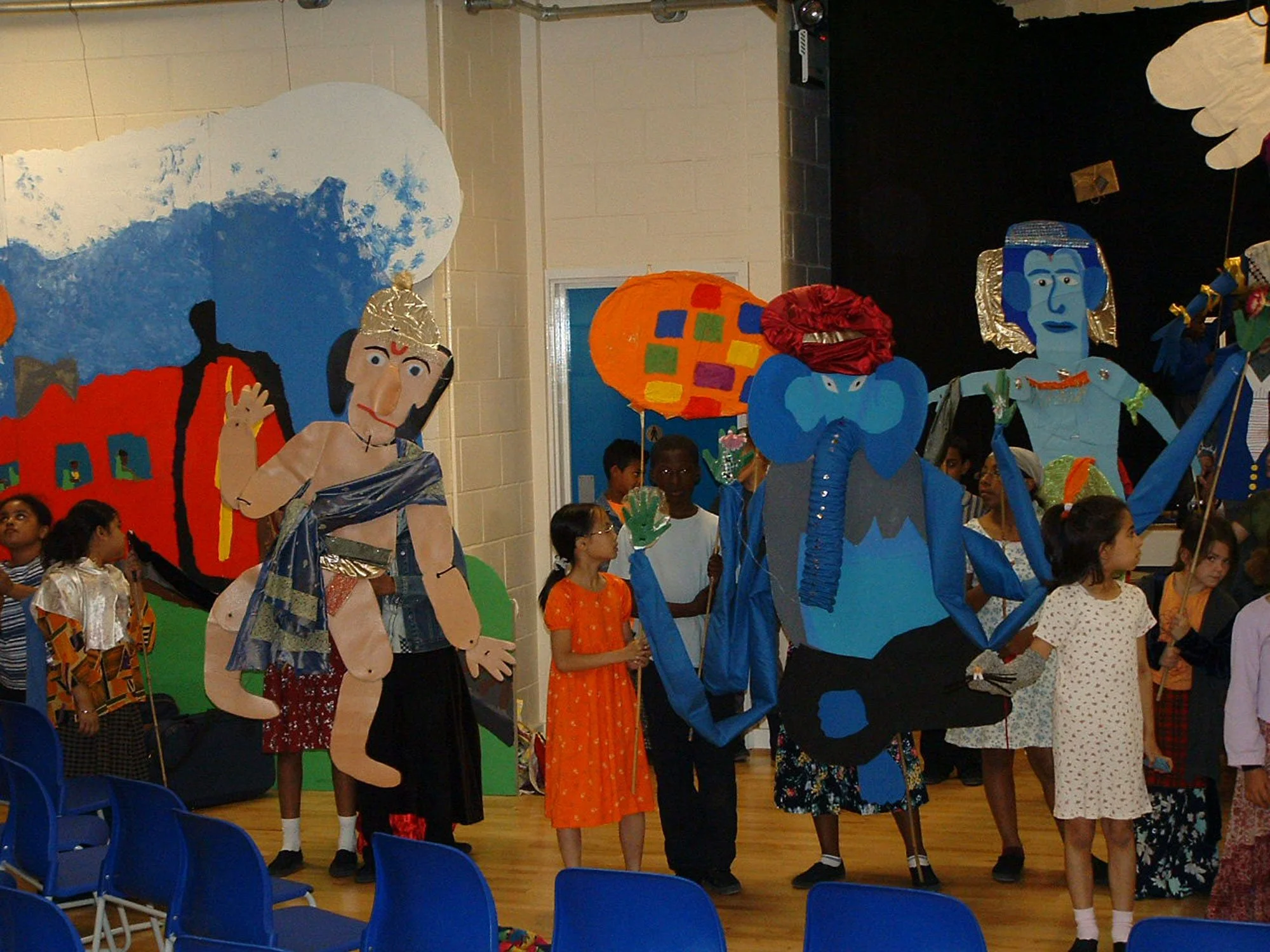 Children participating in a cultural performance with large colorful puppets on stage, some children wearing traditional attire.