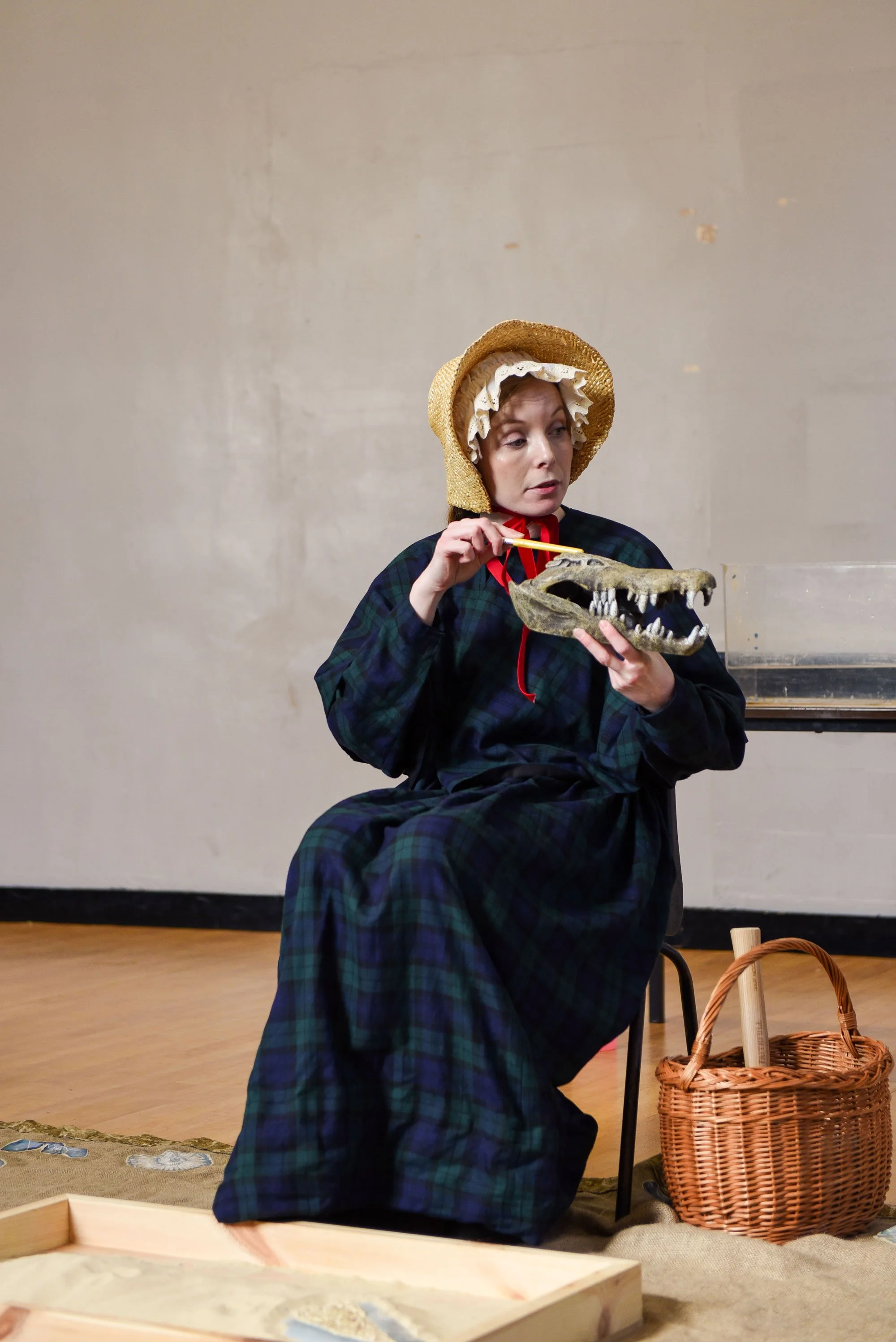 A workshop with an actor playing the part of Mary Anning, dressed in historic clothing, wearing a bonnet, sitting on a chair, holding a dinosaur skull and using a pointer to examine its teeth. There is a wicker basket and a sand tray on the floor.