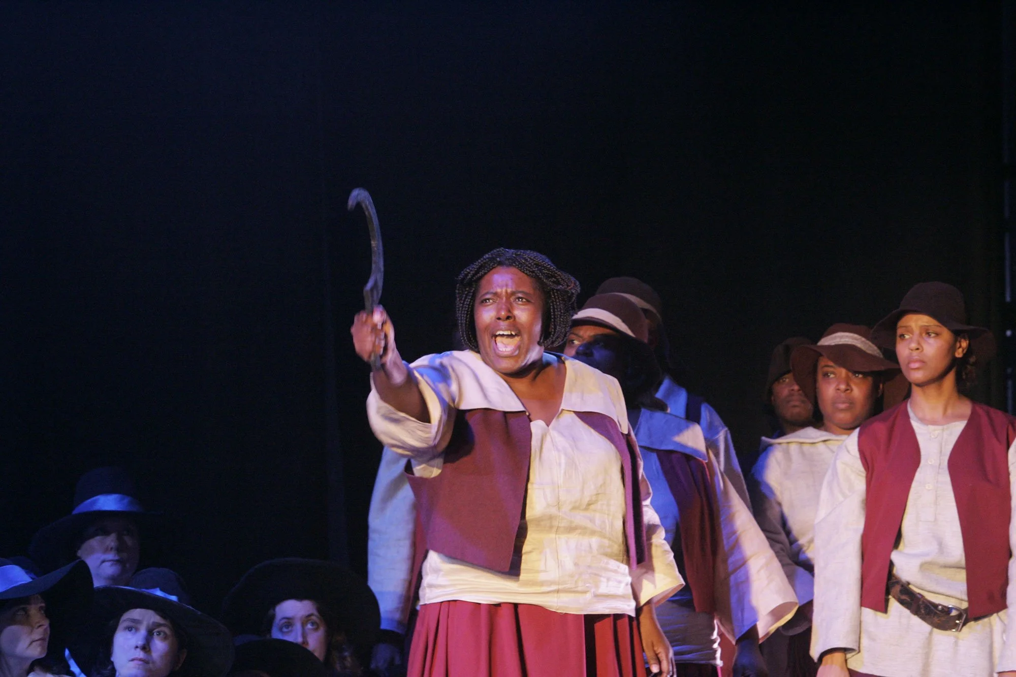 A woman is angrily shouting and holding a hook-shaped object, surrounded by other women who appear attentive or concerned, in a theatrical performance."