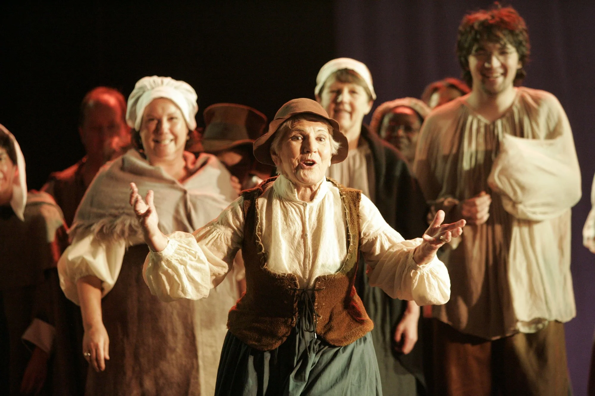 A group of actors in historical costumes performing on stage, with an elderly woman in the foreground gesturing while speaking.