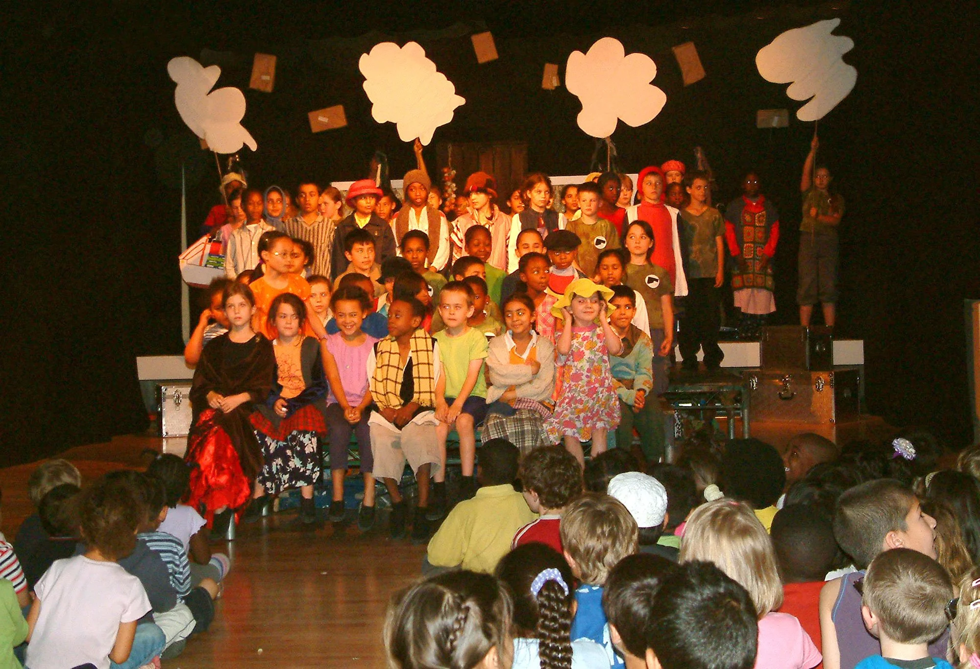 Children on stage during school play, dressed in costumes, with large white cloud cutouts overhead, and children seated in audience watching.