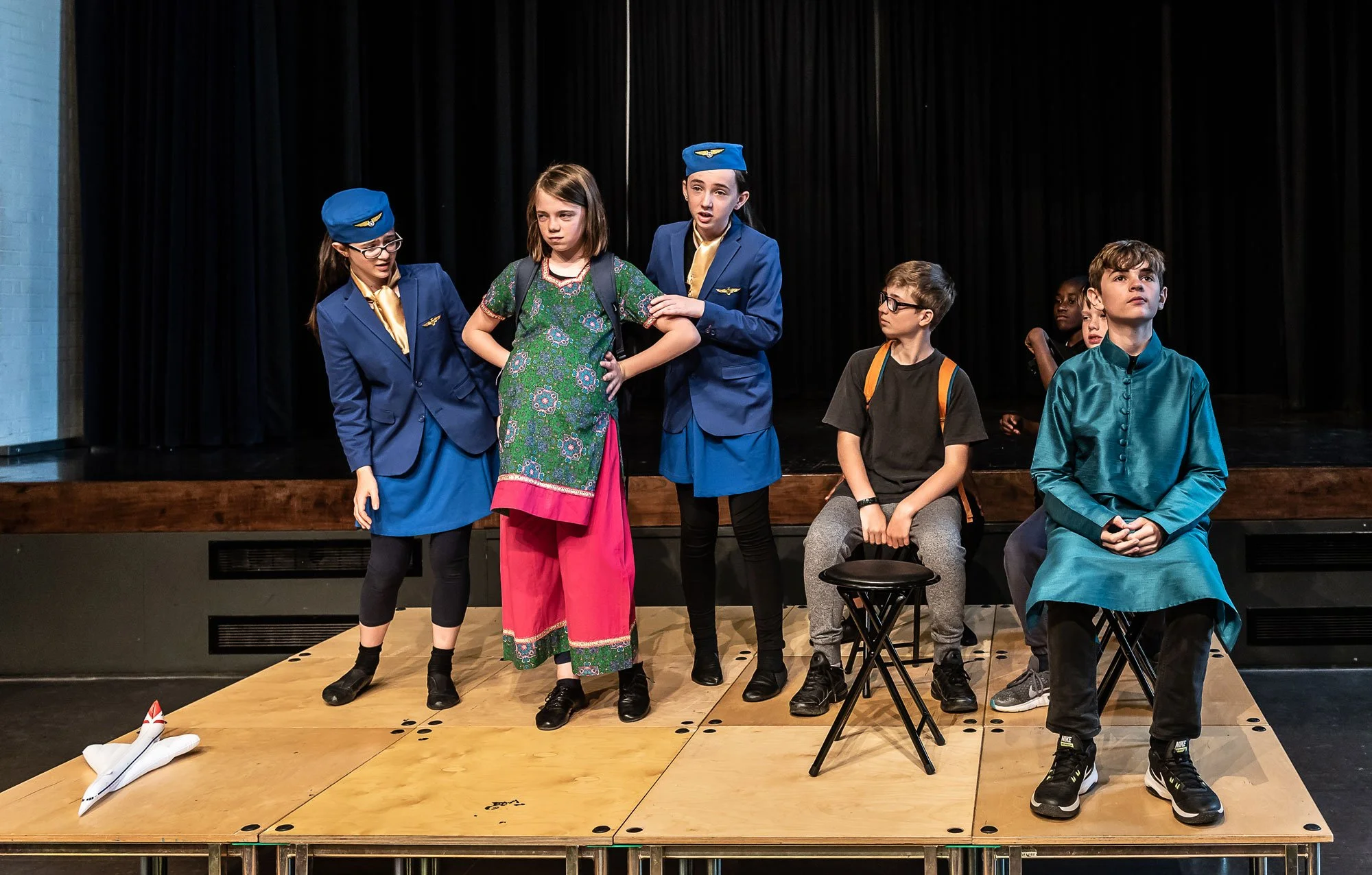 Children performing a school play standing on stage with a black curtain background, some children in pilot costumes, one girl in traditional dress, and two children sitting on stools and chairs.