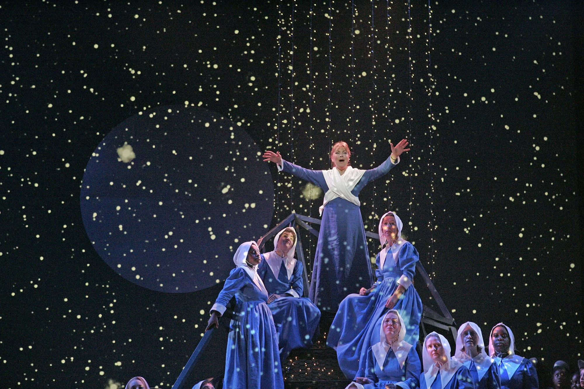 Stage scene from a theatrical production depicting a woman in a blue dress and white shawl standing with arms outstretched on a platform, surrounded by women in blue dresses and white head coverings, with a backdrop of falling snow and a large moon.
