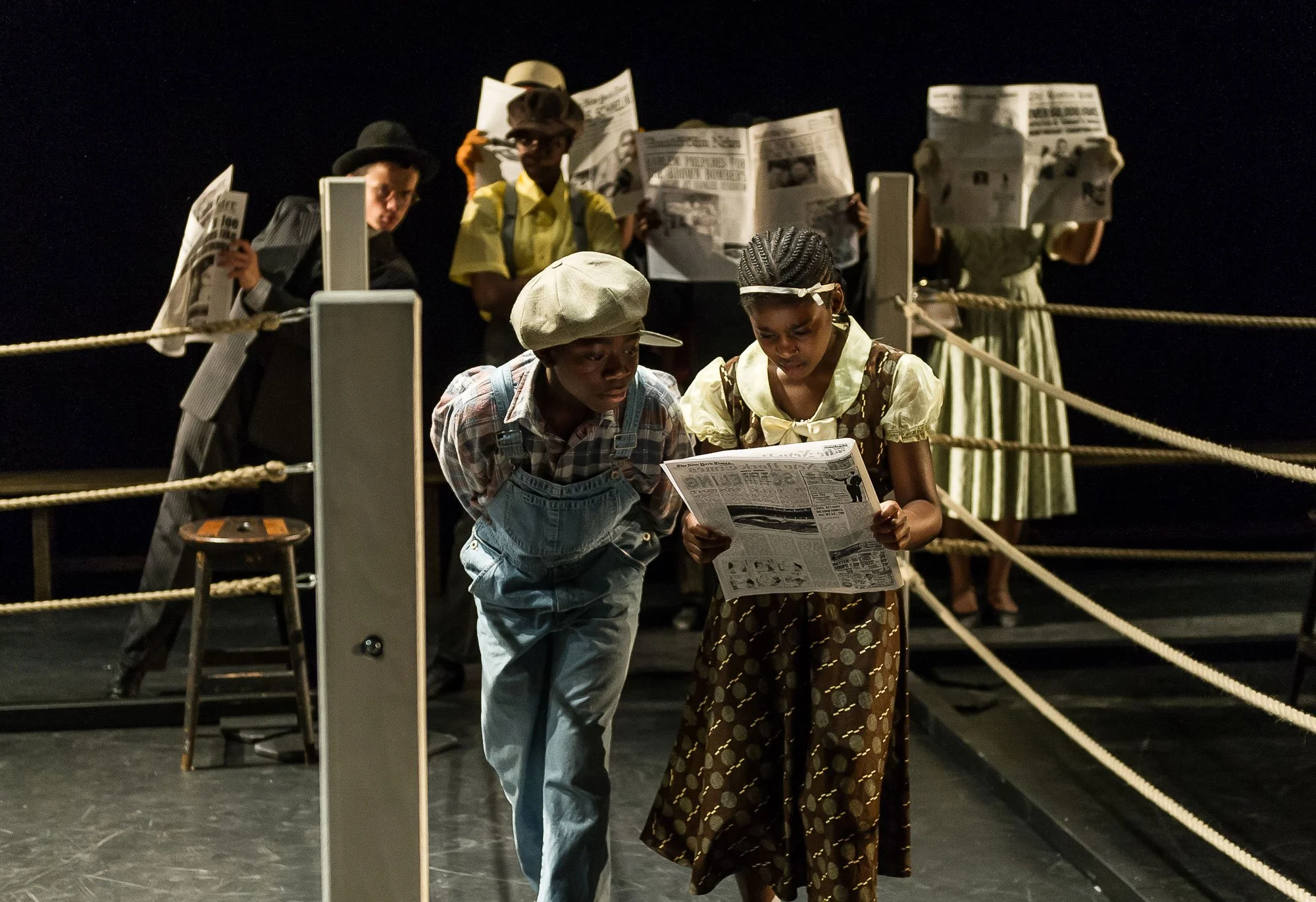 Theater scene with six actors in period costumes, reading newspapers, on a stage with ropes as barriers, dim lighting.