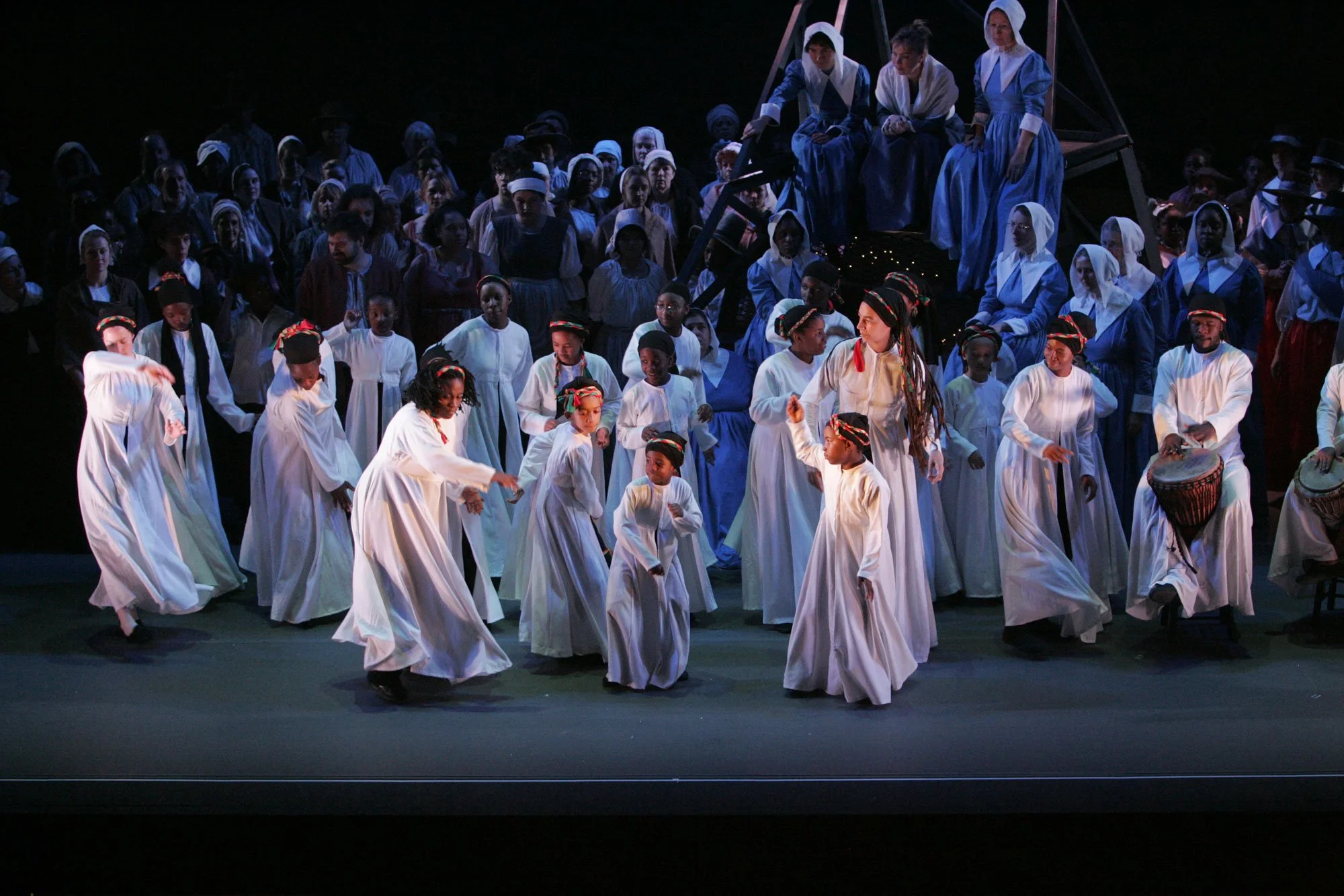 Performers dressed in period costumes dancing and playing drums on stage during a theatrical performance with a large cast in the background.