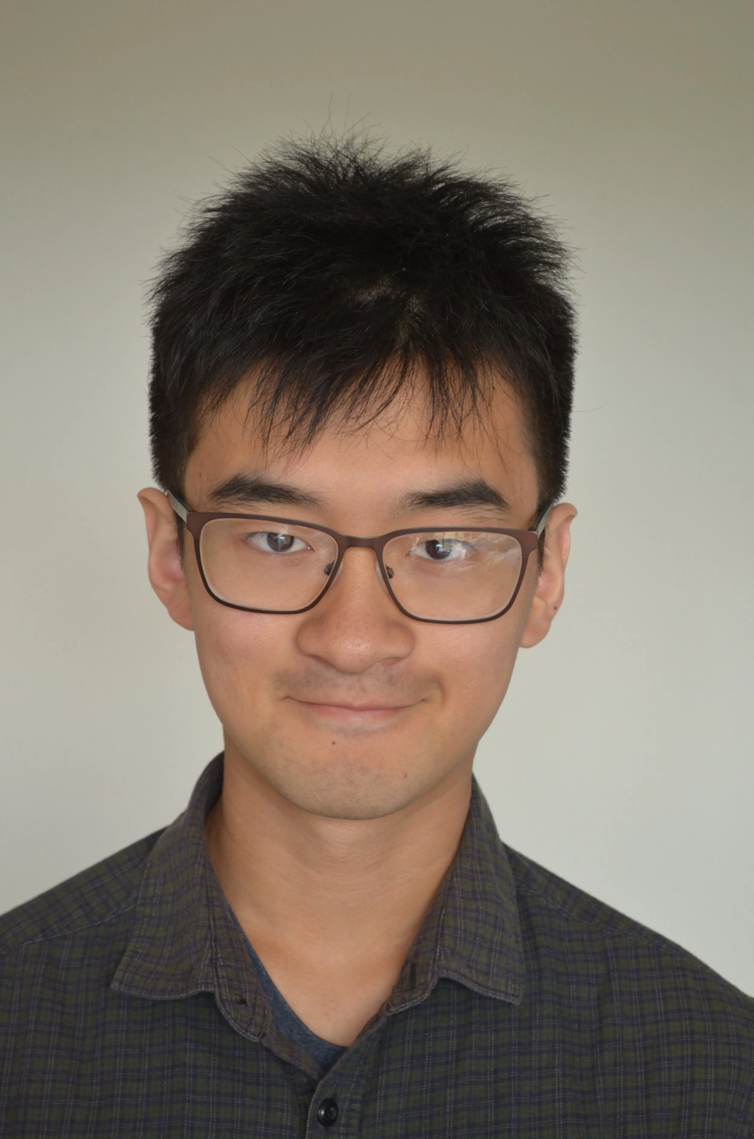 A young man with glasses, short black hair, and a slight smile, wearing a dark green checkered shirt, standing against a plain light background.