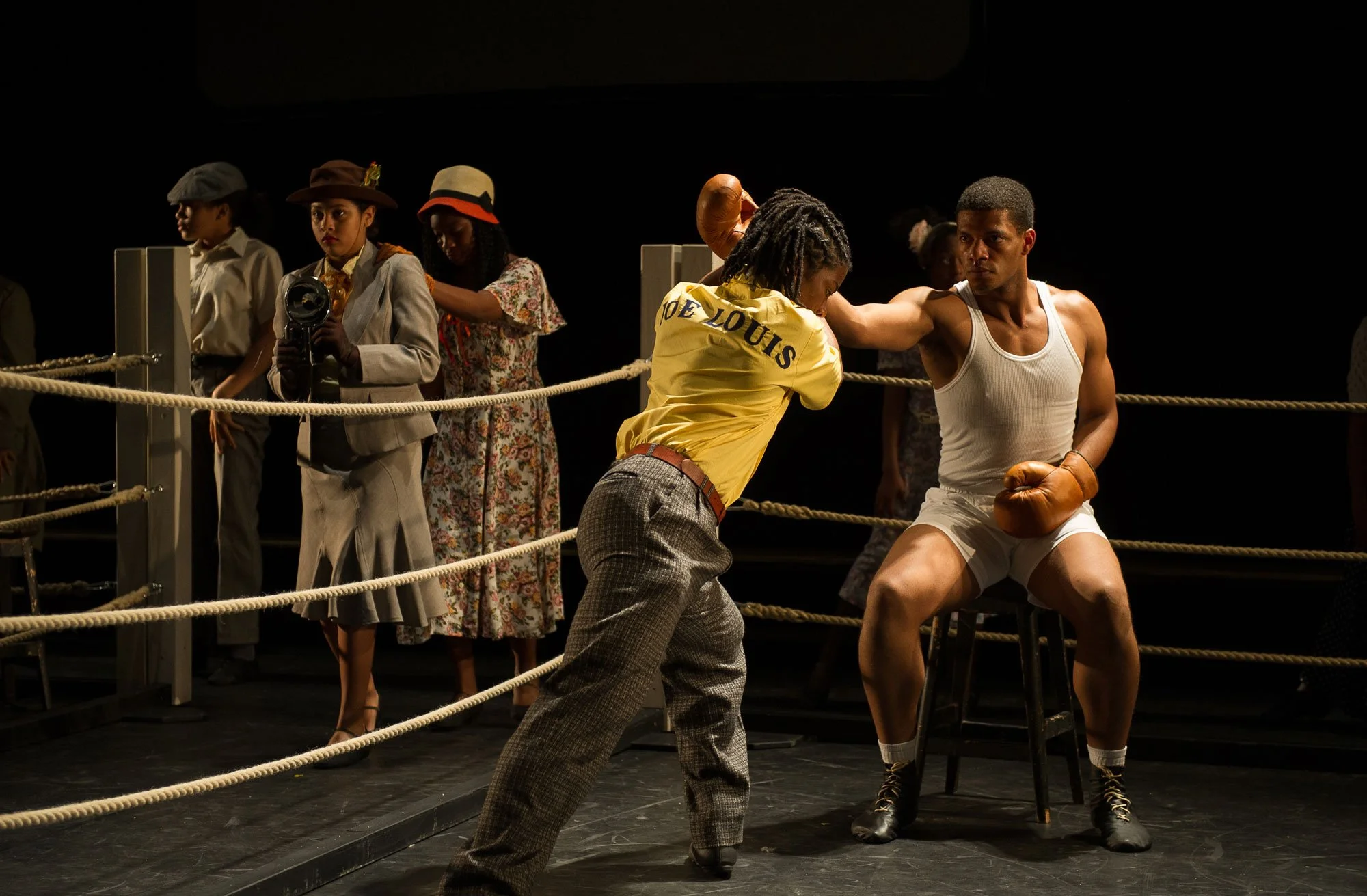 A scene from a theatrical boxing match with actors, including one in a vintage suit and another in athletic wear, boxing in a ring while others watch.