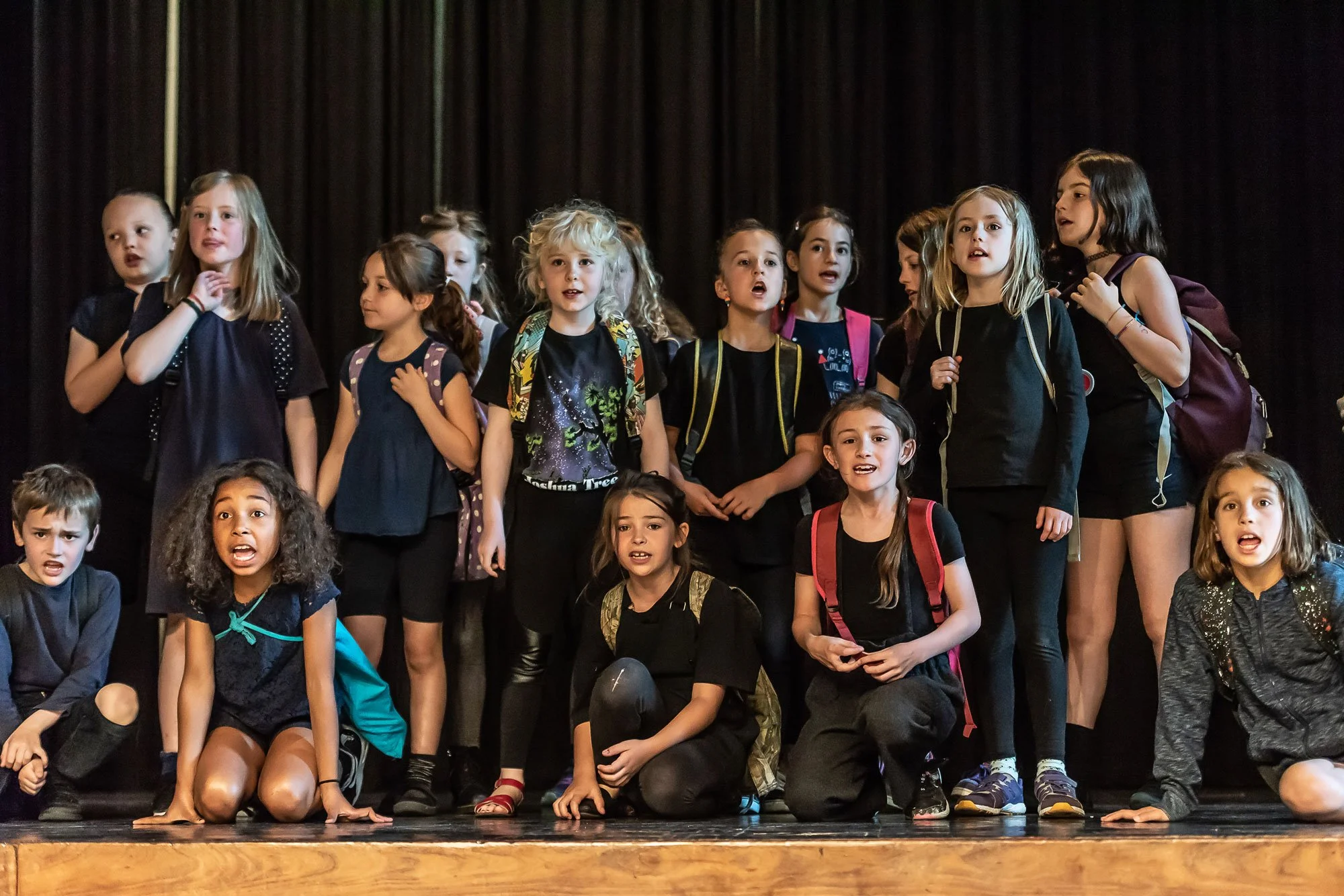 A group of children on stage, some standing and some kneeling, singing or performing with a dark curtain backdrop.