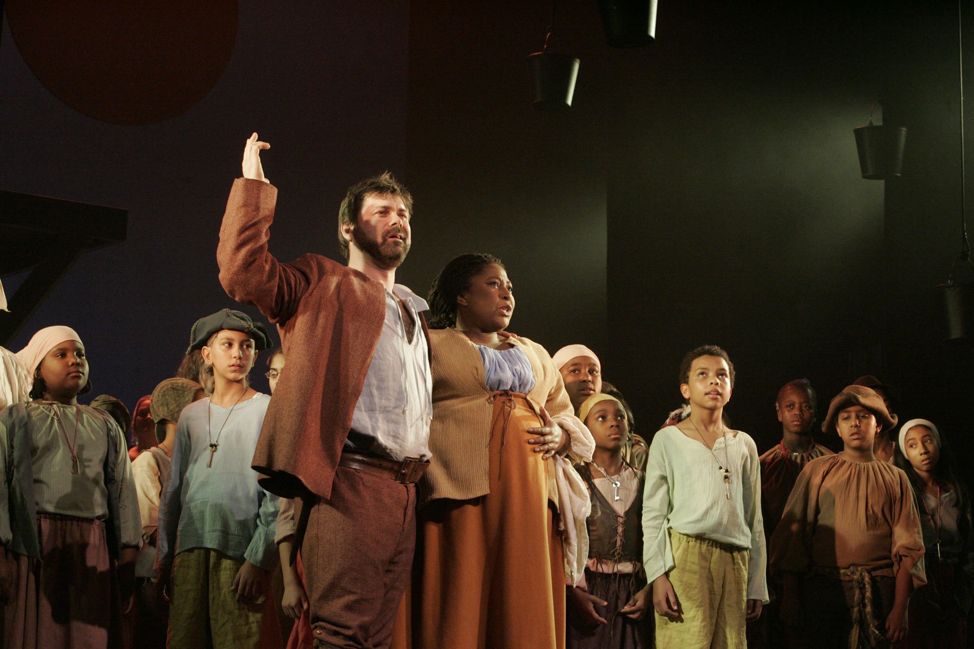 A group of people on stage, with some raising their right hands, performing in a theatrical production. The scene features diverse characters in period costumes, with dark background and stage lighting.