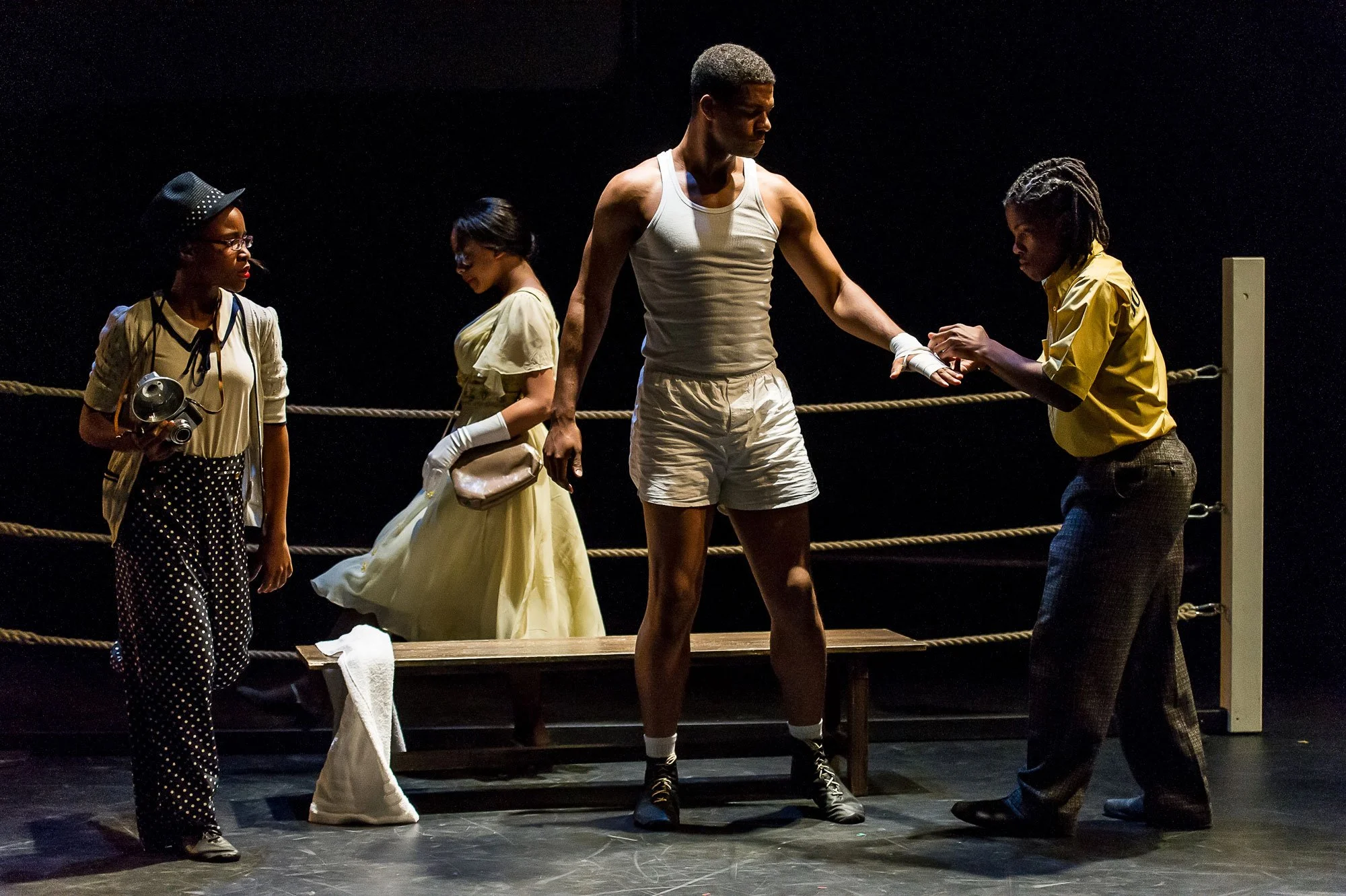 The image depicts a theatrical scene with five actors on stage. The central figure is dressed in white athletic shorts and a tank top, reaching out and holding hands with a woman in a yellow shirt and dark pants. Two women in vintage dresses and gloves stand in the background, and another woman in polka-dot pants and glasses holds a camera, observing the scene.