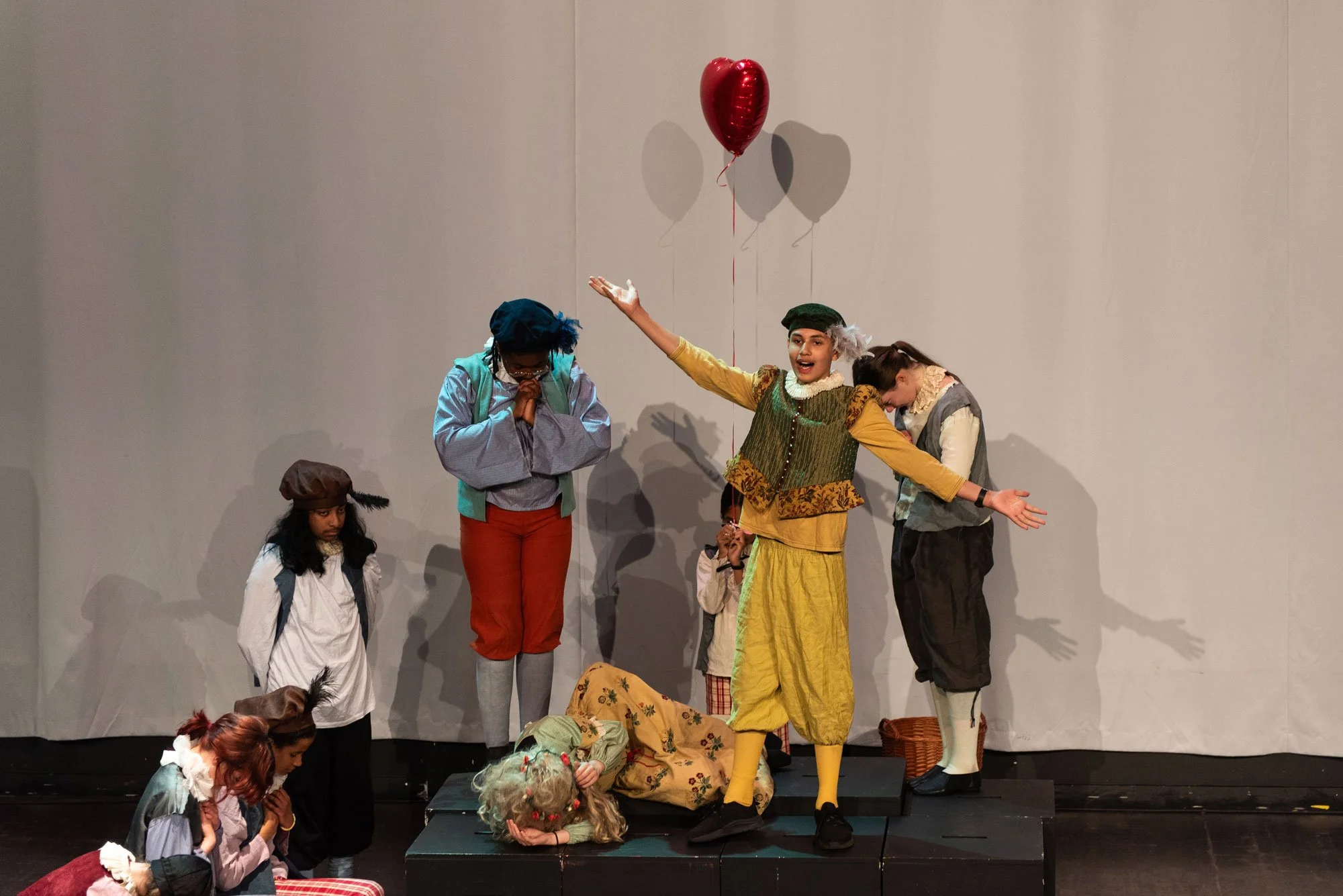 A group of children performing a theatrical play on stage, dressed in colourful costumes with props such as a red heart-shaped balloon, a basket, and fake animals, against a plain white background.