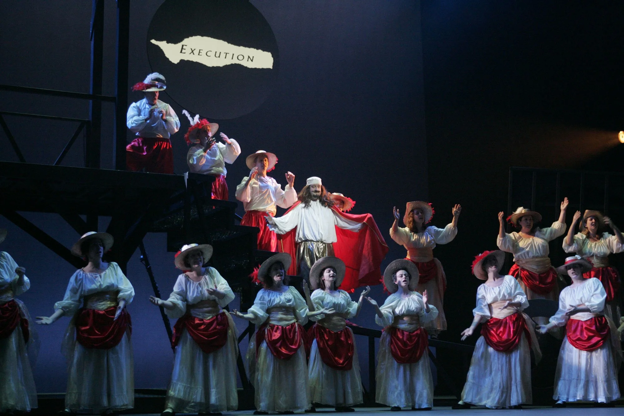 A theatrical stage performance featuring people dressed in historical costumes, with some wearing large hats and red sashes, on a multi-level set. The background displays a sign that reads 'Execution'.
