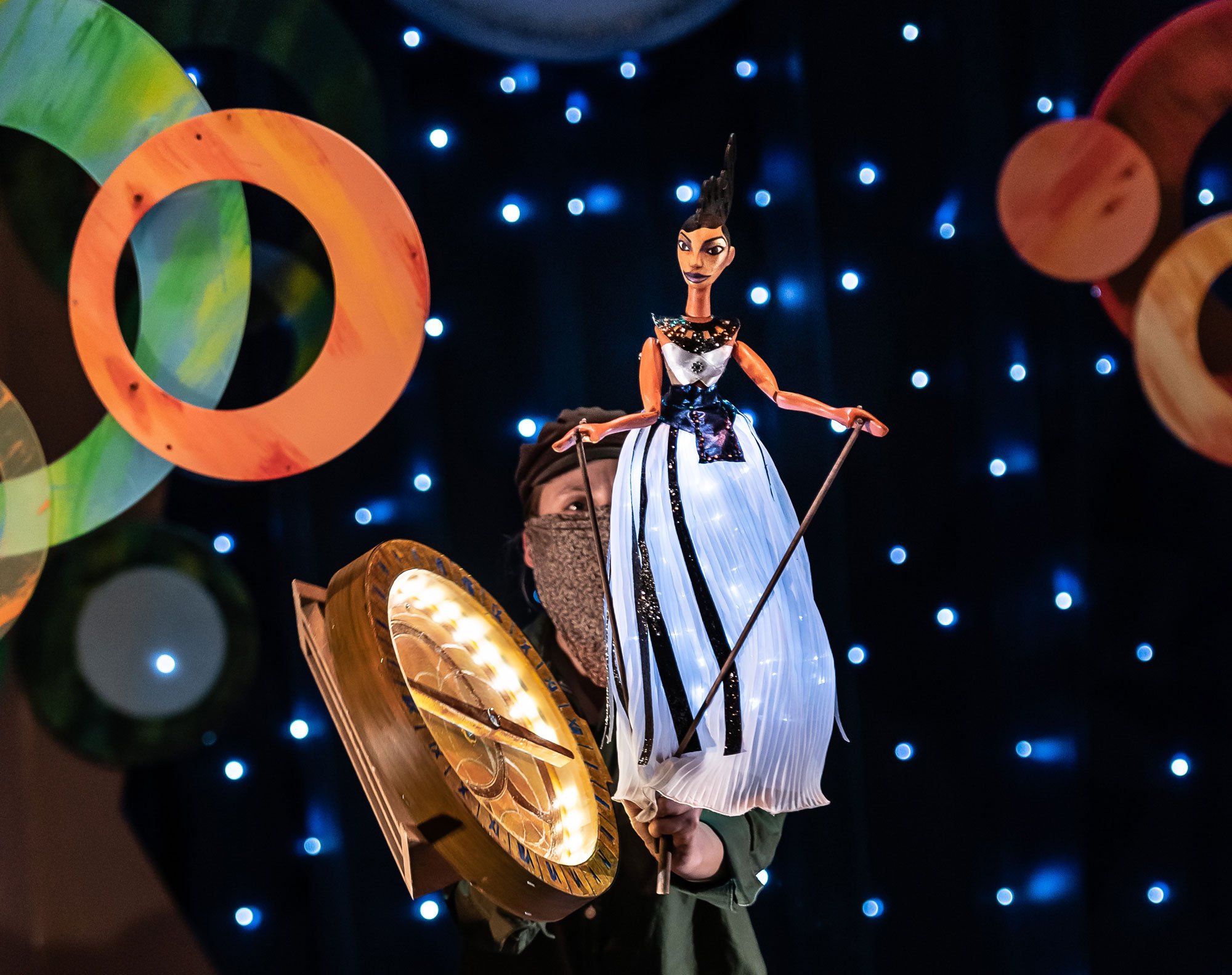 A puppeteer holding a marionette puppet of Maria Sibylla Merian dressed in a white gown, against a background of blue lights and colourful circles.