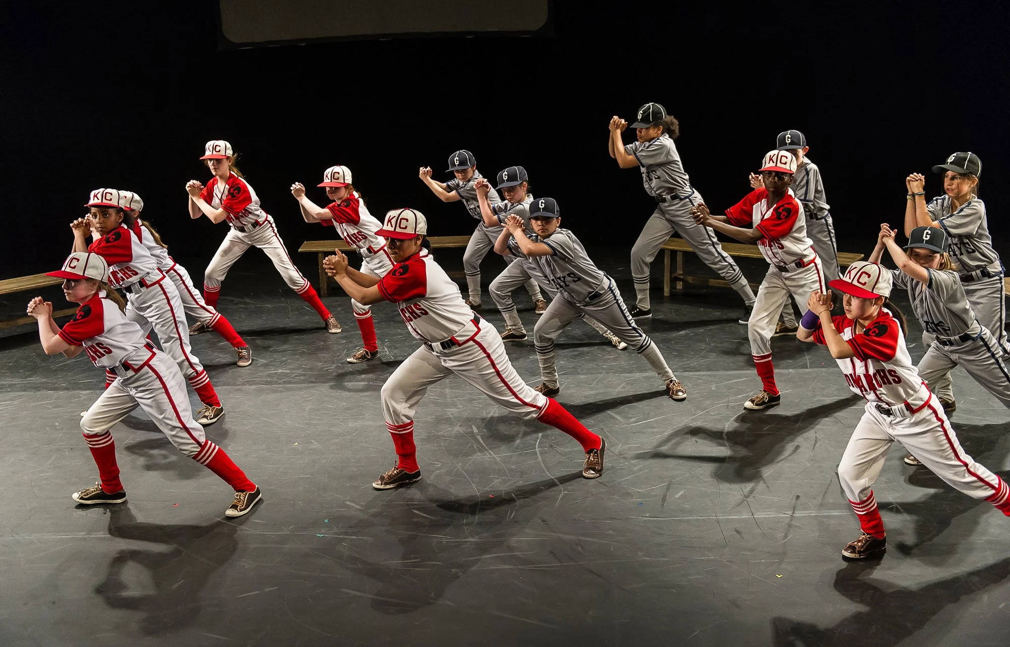 Young girls and boys in baseball uniforms performing a choreographed dance or pose on a stage with a black background.