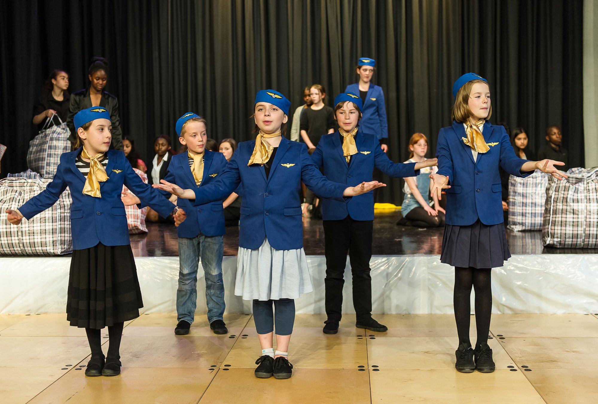 Children dressed as airline flight attendants perform on stage, with some seated in the background and others standing in front.